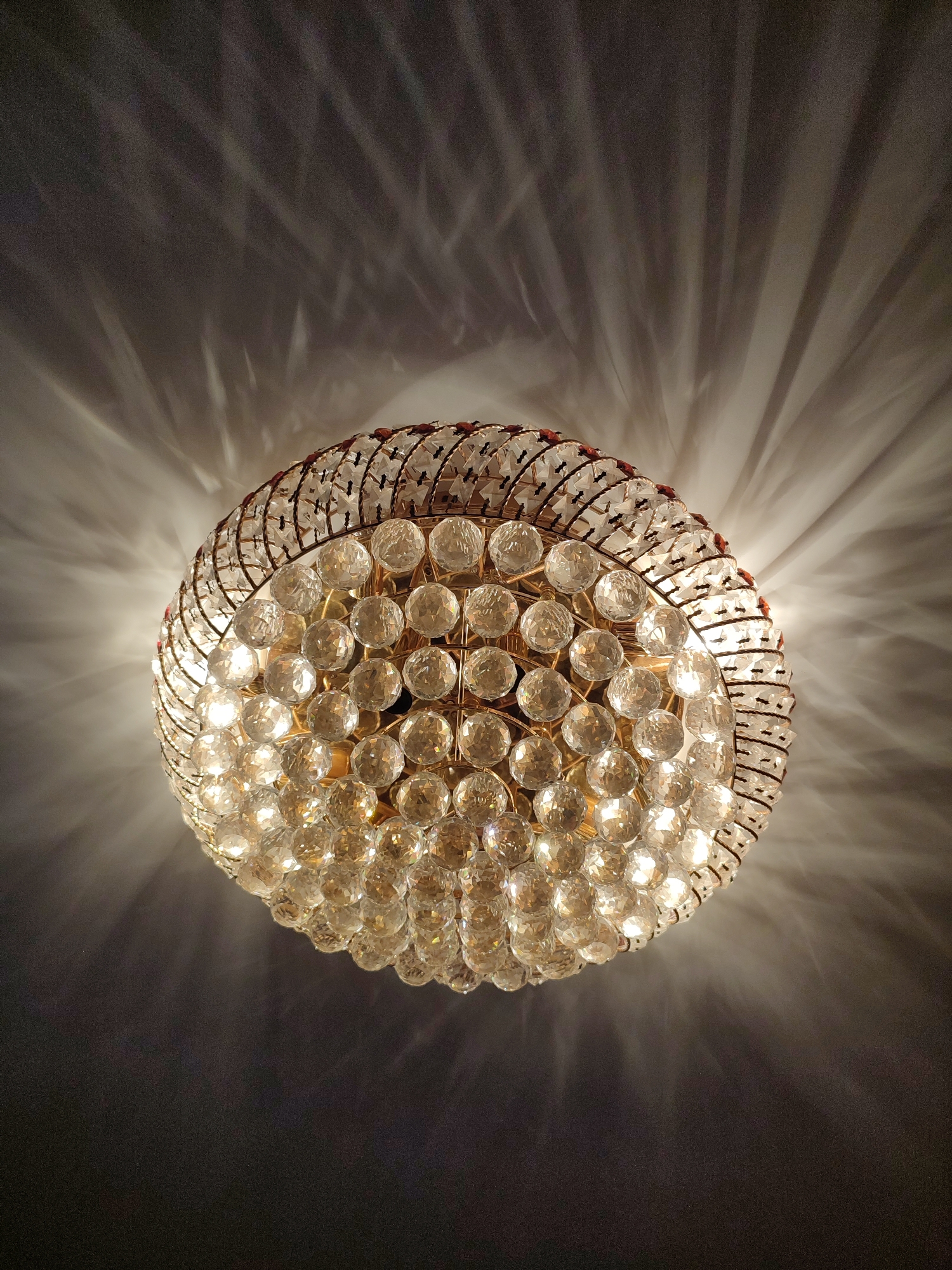 The image shows a close-up, low-angle shot of a chandelier hanging from a ceiling. The chandelier is round and appears to be made of brass or a similar metallic material, with numerous clear, faceted crystal spheres arranged in a spherical pattern. These spheres catch and refract the light, creating a shimmering effect and casting intricate patterns of light and shadow onto the ceiling above. The light source within the chandelier is visible, illuminating the crystals and casting bright highlights. The ceiling is dark, with the light from the chandelier creating dramatic, radiating beams and abstract shapes. The overall impression is one of elegance and luxury, with the play of light and shadow adding a dramatic and somewhat ethereal quality to the scene. There is no text visible in the image, and no indication of the time of day or weather conditions. The setting appears to be indoors, likely in a room with a high ceiling, given the way the light is projected. No people are visible.