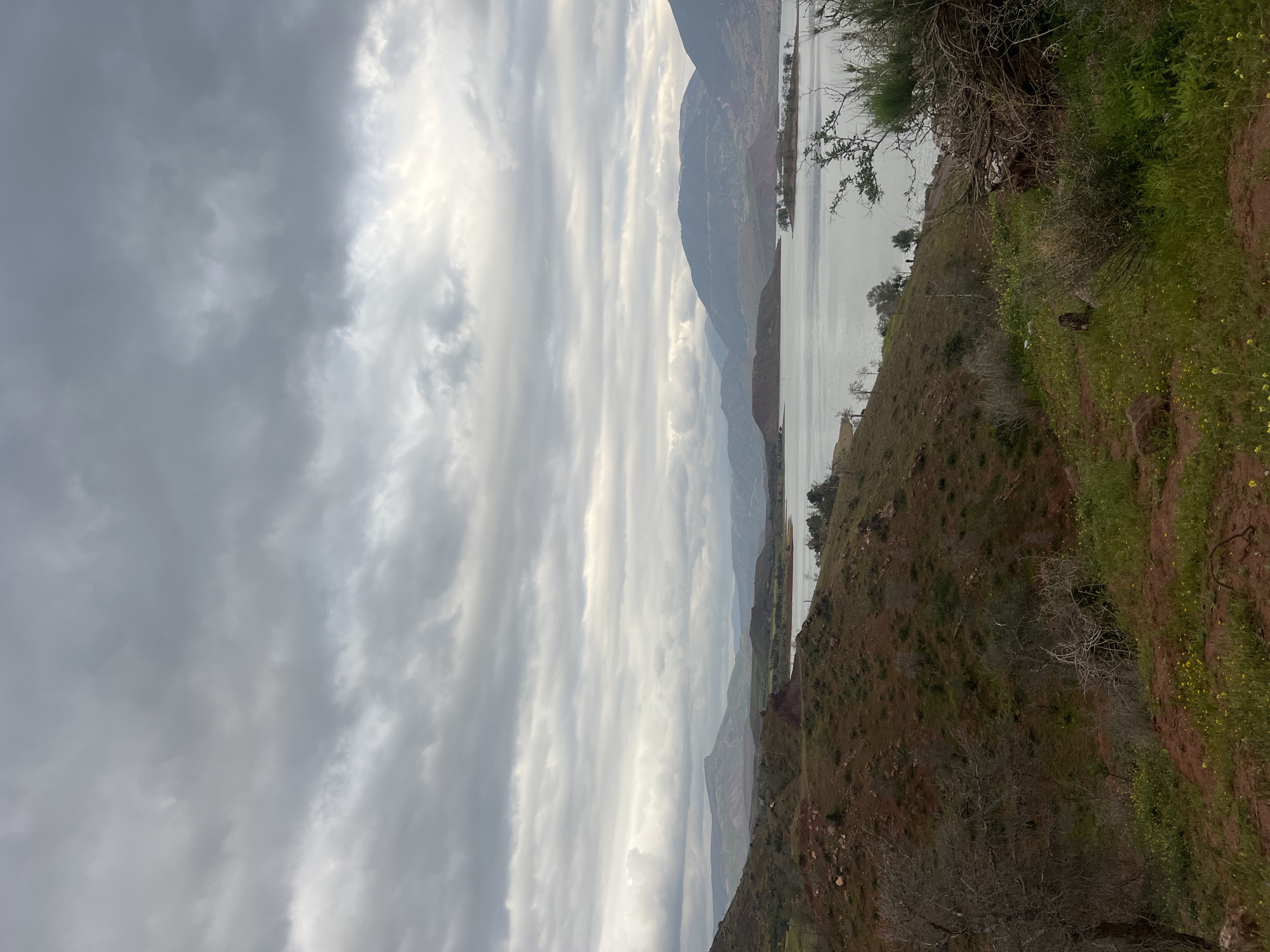 The image captures a landscape near Bin El Ouidane, Morocco. The scene presents a body of water, likely part of the Bin El Ouidane lake, bordered by hills and mountains. Overcast skies with layered clouds dominate the upper portion of the image.
The landscape features grassy slopes with patches of exposed soil and sparse vegetation, including some yellow wildflowers. The foreground consists of a hillside descending toward the water.