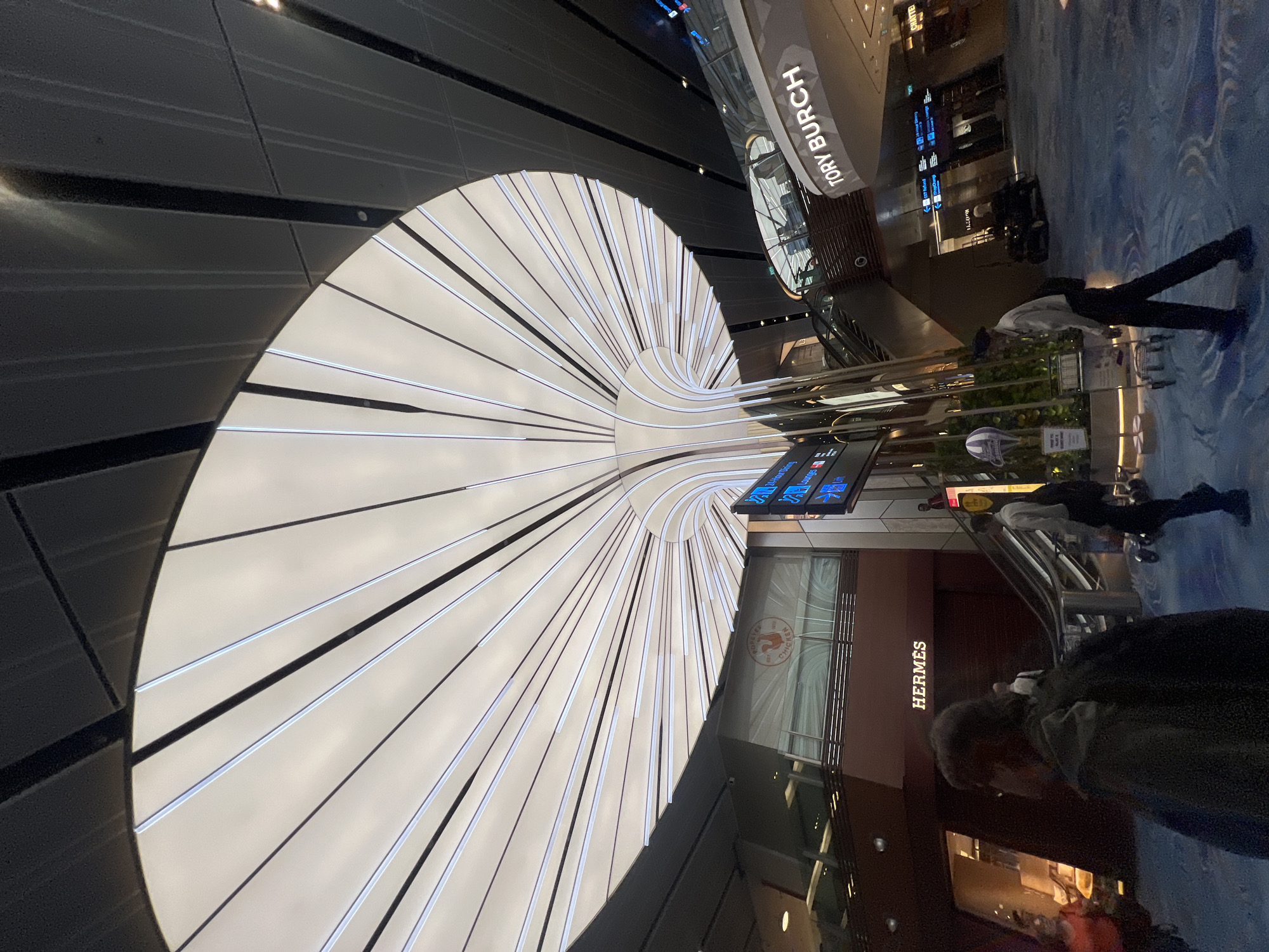 The image depicts the interior of Jewel Changi Airport in Singapore. The scene is characterized by a striking, large, petal-like ceiling structure with internal lighting. Below this, a central pillar curves upwards, supporting the ceiling. To the left, a store with the "HERMES" logo is visible, and above it, a "POPEYE'S CHICKEN" sign. To the right, a sign for "TORY BURCH" is prominently displayed. Several individuals are walking around the airport's spacious, blue-carpeted floor. An escalator is visible in the background, leading to a higher level. Information boards displaying flight details or directions are also present. The overall environment suggests a bustling airport terminal with various retail outlets and amenities.