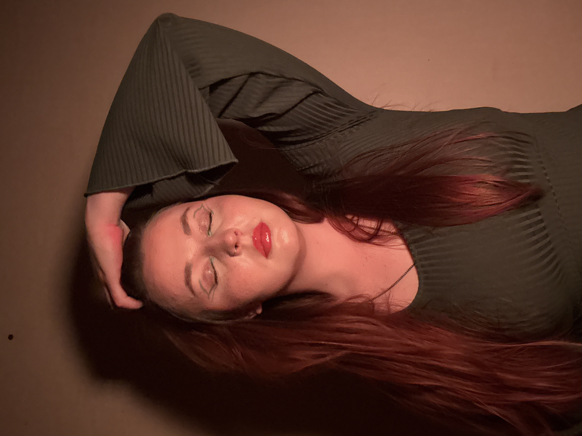 A young woman lies on her back on a flat, possibly textured, surface that appears to be a mottled brown color. Her head is tilted slightly back and to her right. Her long, red hair is spread out around her head and neck. She has fair skin, closed eyes with what appears to be light green eye makeup, and bright red lipstick. She wears a dark green, long-sleeved dress with a ribbed texture and flared sleeves. Her right arm is bent above her head, with her hand touching her hair. A thin chain necklace is visible around her neck. The lighting is soft and uneven, with shadows falling across her face and body. There is a dark spot on the surface near her left hand. The image provides no indication of Izhevsk, Russia, or any city.