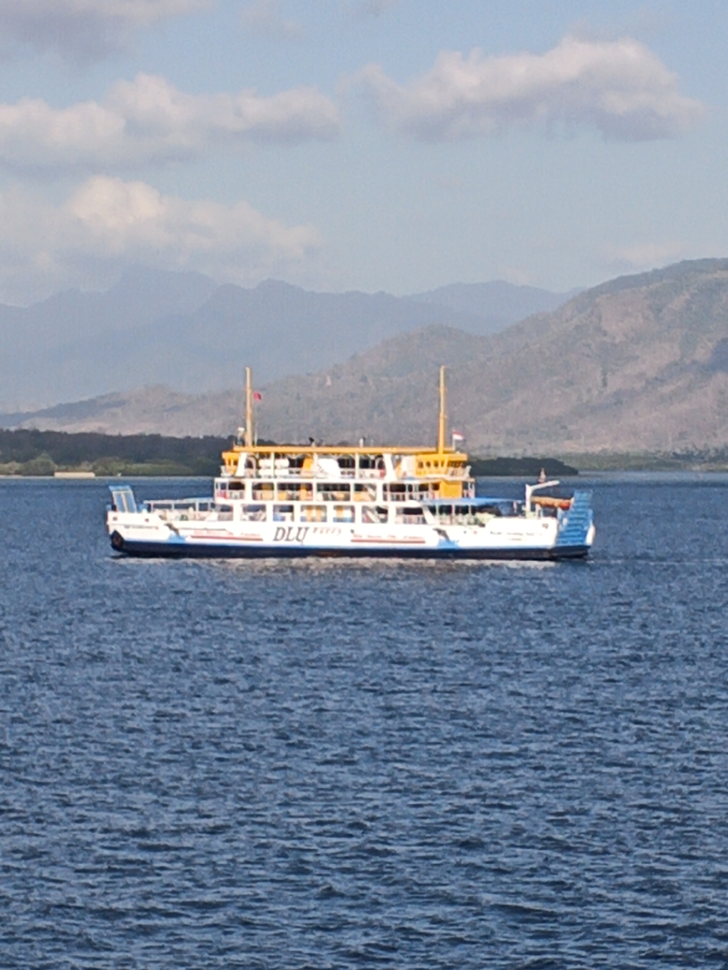A large, white passenger ferry with yellow accents and blue trim sails across a body of water. The ferry has multiple decks visible, with windows lining each level. The name "DLU Express" is clearly visible in blue lettering on the side of the vessel. Two masts rise from the top deck, one with a red flag. The ferry is positioned in the center of the frame, with the water occupying the lower half of the image. In the background, a low, tree-covered coastline is visible, beyond which are rolling, mountainous hills under a bright, cloudy sky. The water is a deep blue, with small ripples on the surface. The overall scene suggests a sunny day in a coastal area of Indonesia.