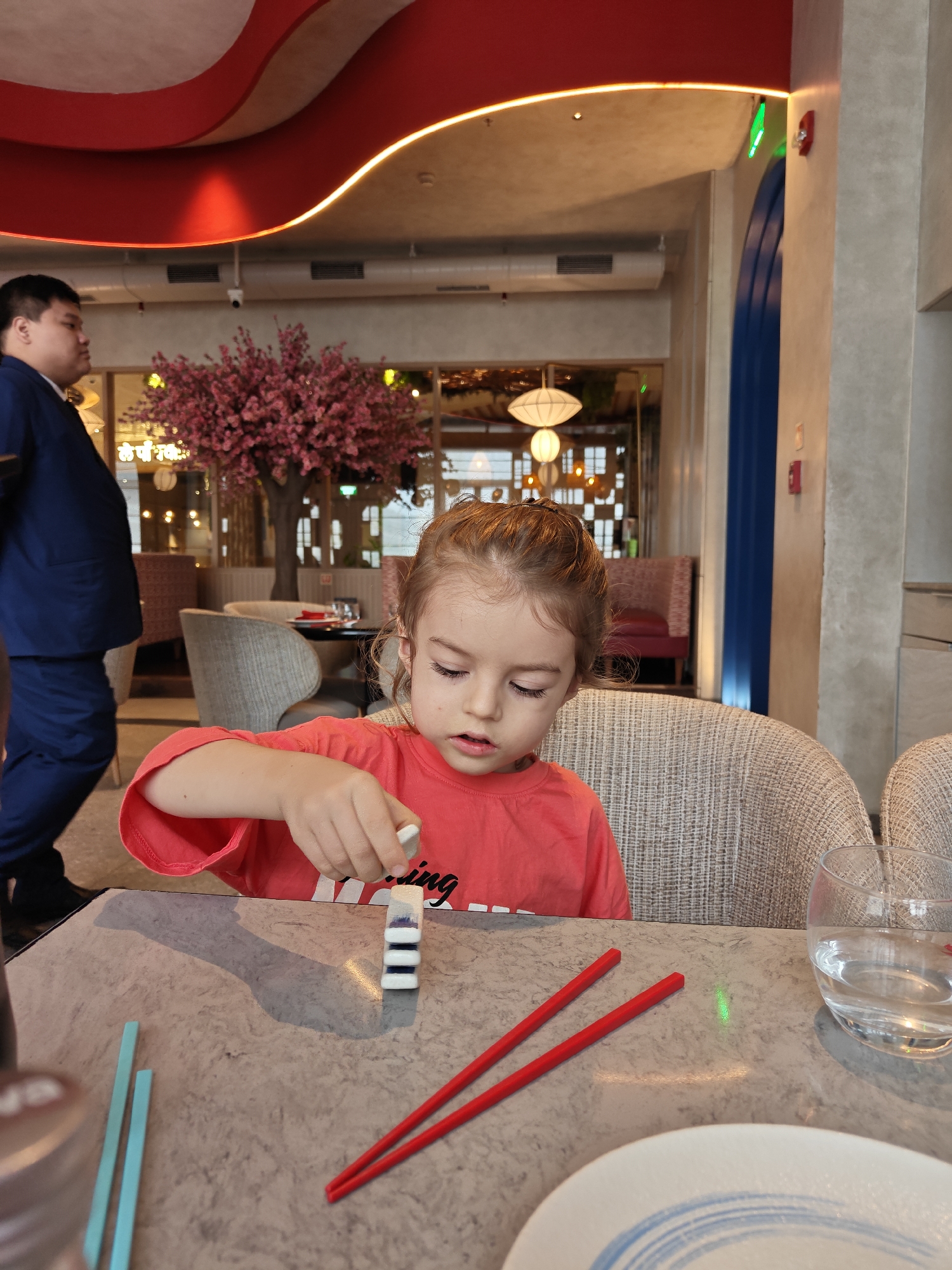 A young girl with brown hair pulled back in a bun, wearing a bright coral t-shirt, is seated at a table, focused on building a small tower of what appear to be dominoes. Her right hand is extended, placing a white and blue domino piece onto the stack. In front of her are red chopsticks and two light blue rectangular objects, possibly placemats or coasters. A glass of water sits to her right, and a plate with blue concentric circles is partially visible in the foreground. The table surface is a mottled grey. Behind the girl, a man in a dark blue suit is walking away from the camera, with his back turned. The background reveals a restaurant setting with a decorative red ceiling feature, a flowering pink tree, and seating areas with upholstered chairs. The atmosphere suggests a dining establishment. The image does not provide any specific location details for Mumbai, India.