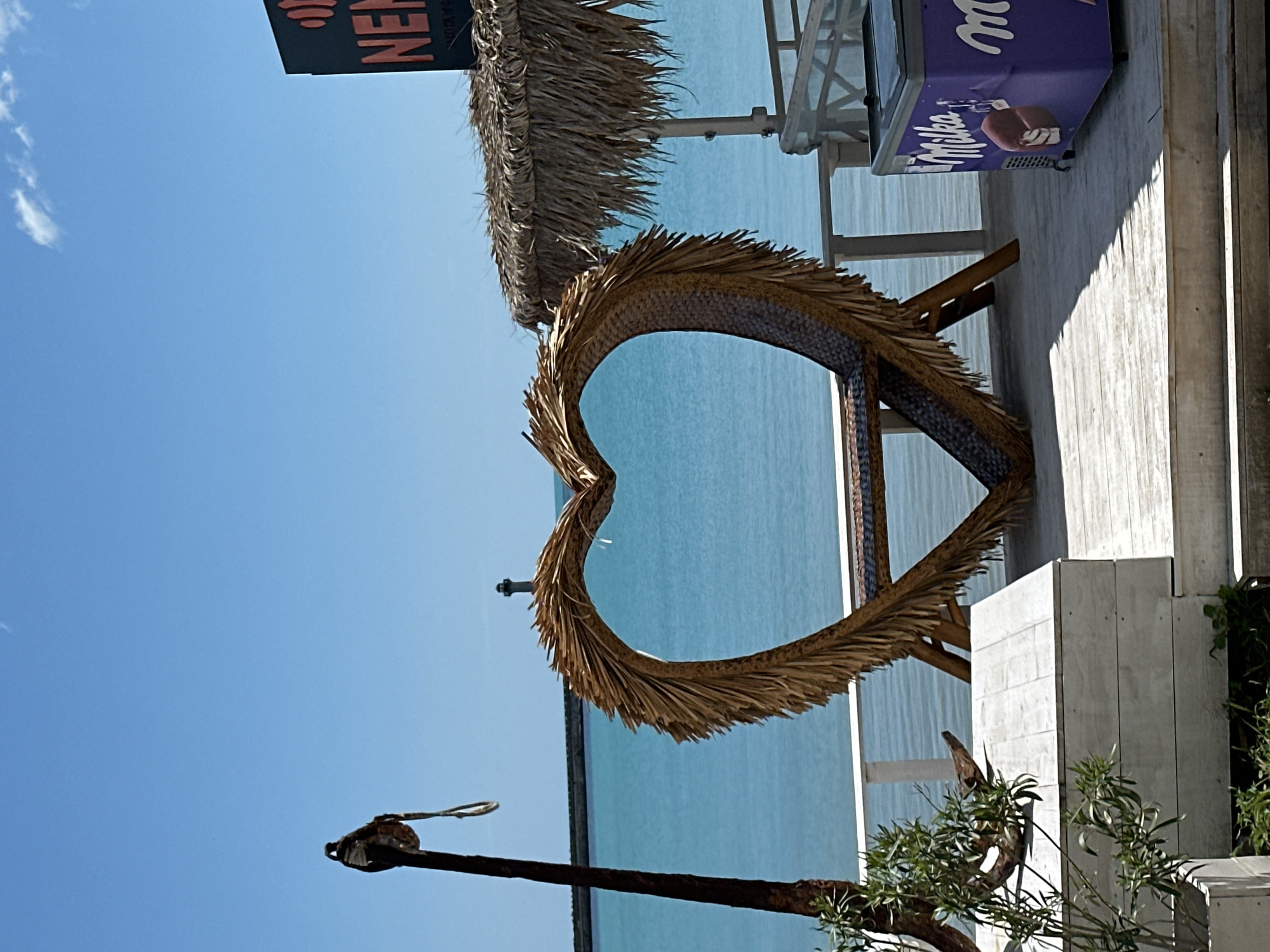 The image shows a view of the Black Sea in Varna, Bulgaria. In the foreground is a large heart-shaped structure made of straw-like material, positioned on a wooden platform. To the right of the heart is a white wooden structure, possibly part of a building, and a purple Milka ice cream freezer. In the background, the blue water of the Black Sea stretches to the horizon under a clear blue sky. To the left, an anchor is visible, mounted on a rusty pole. A small sign is visible above the heart, advertising "New Beach". The scene conveys a beachside setting in Varna.