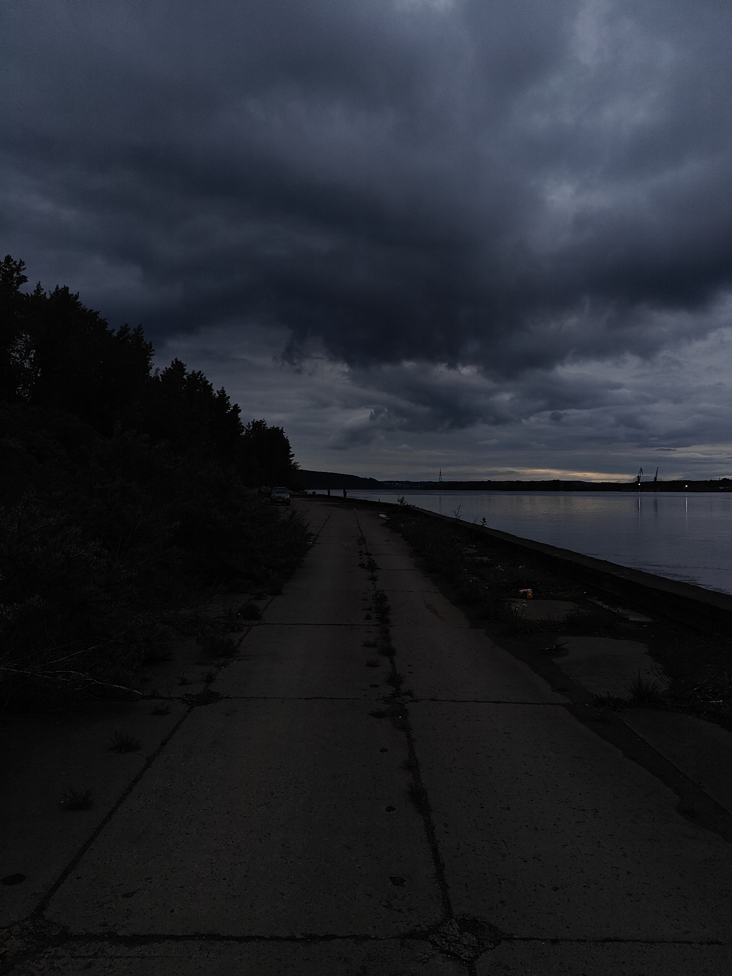 The image shows a concrete pathway leading into the distance alongside a body of water. Dark, heavy storm clouds dominate the sky. To the left of the path is a dark, wooded area. To the right, the water's edge is lined with concrete blocks. In the distance across the water, structures resembling cranes are visible. The setting appears to be dusk or night. The location is likely Chaikovsky, Russia, based on the prompt. No people are visible.