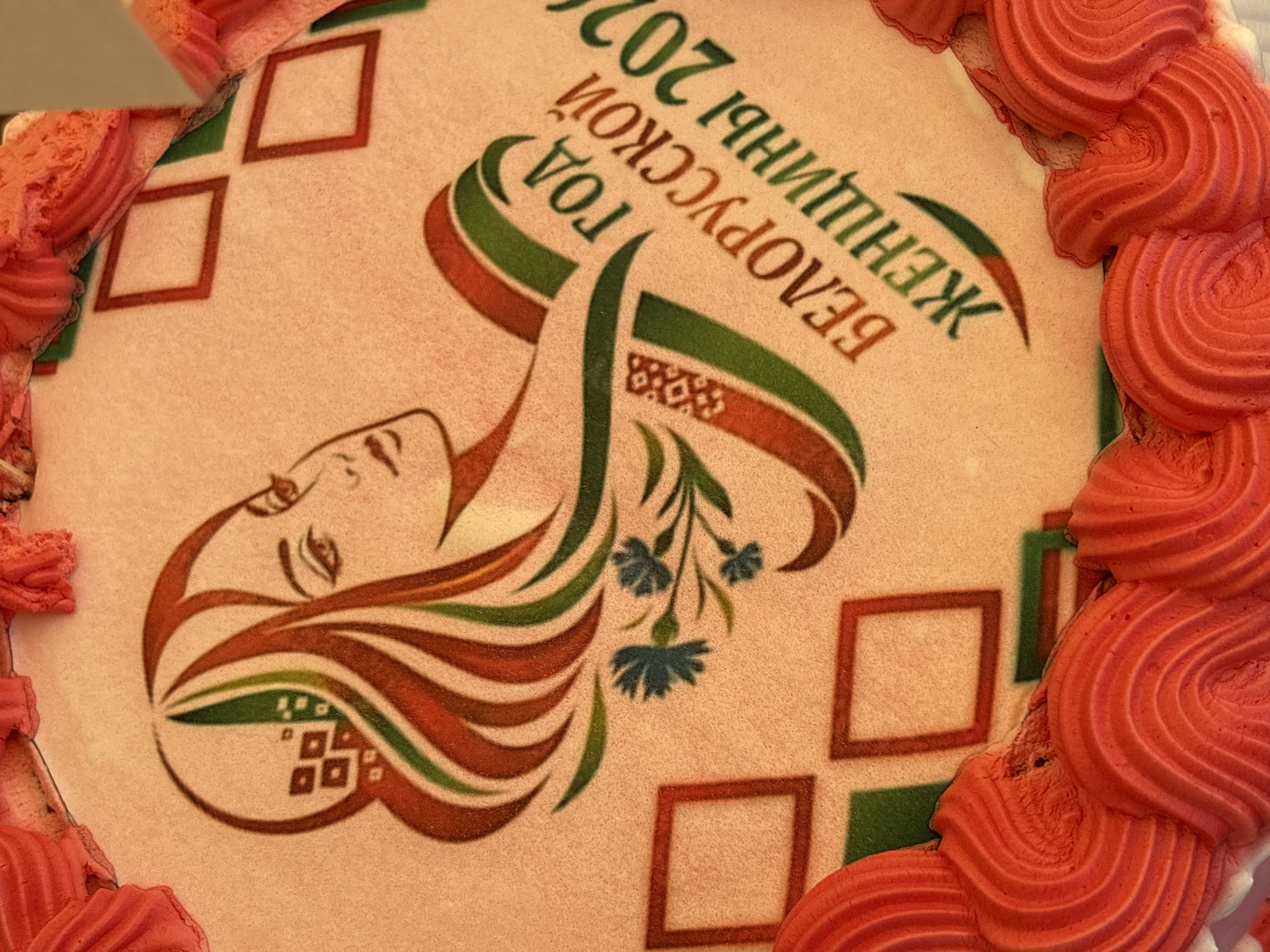 This is a close-up, top-down view of a cake, likely decorated for a celebration. The cake has an edible image on its surface depicting a stylized profile of a woman with long, flowing hair in shades of red, green, and brown. Accompanying the portrait is text in Russian: "ГОД БЕЛОРУССКОЙ ЖЕНЩИНЫ 2020", which translates to "Year of Belarusian Women 2020". There are also abstract geometric patterns in red and green squares on the cake's surface, and a small cluster of blue flowers. The edges of the cake are adorned with red frosting piped in a ruffled or wave-like pattern. The image does not provide information about its location in Вяйнянскі сельскі Савет, Belarus.