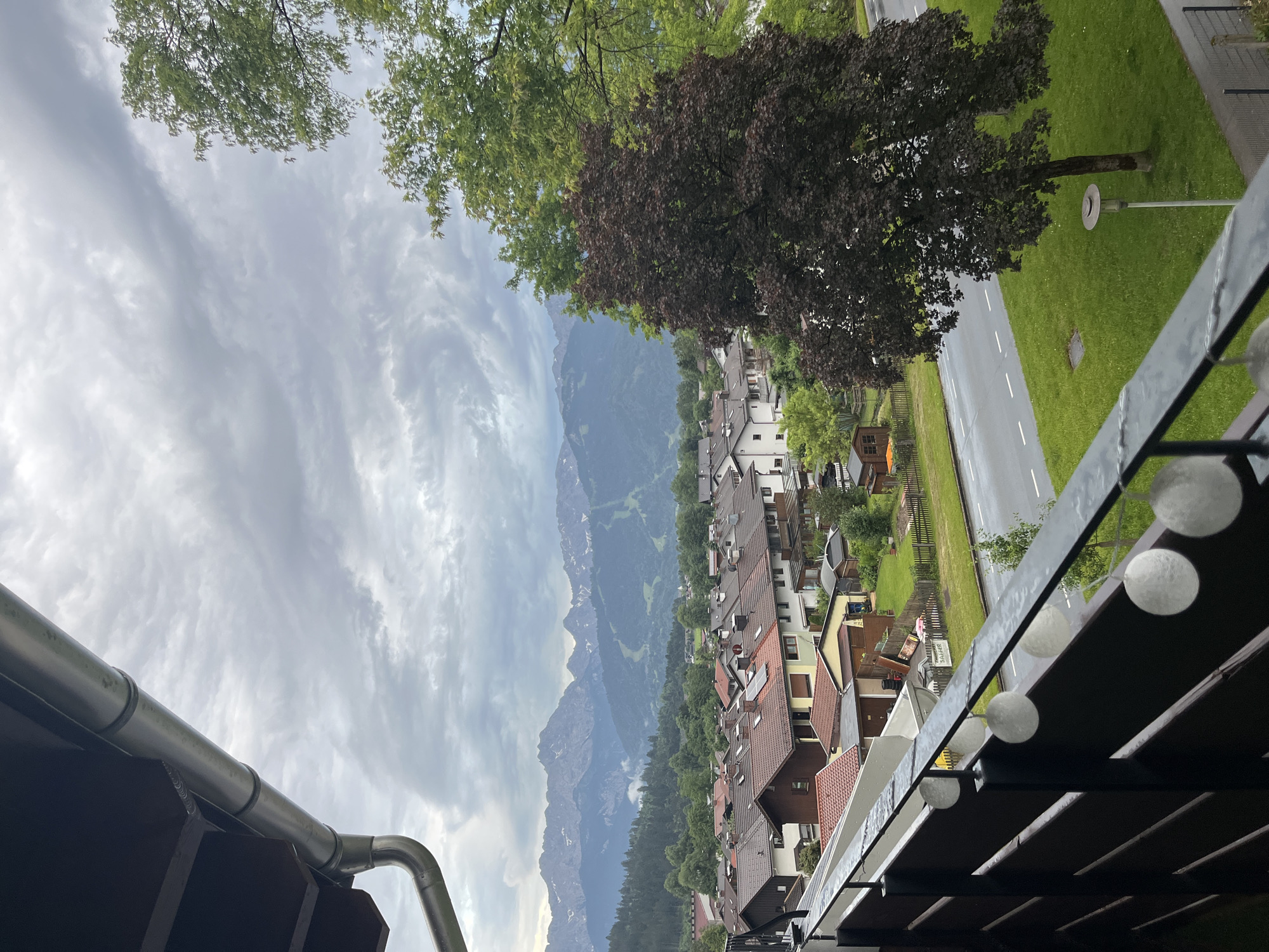 The image presents a cloudy day view from a balcony overlooking a suburban area in Garmisch-Partenkirchen, Germany. In the foreground, a dark wooden balcony railing is visible, adorned with a string of spherical white lights. To the left, a portion of a building's dark brown overhang and a silver gutter system are present.
Beyond the balcony, a street runs horizontally across the mid-ground. Adjacent to the street is a well-maintained green lawn, featuring a single tree with dark reddish-brown foliage and a modern lamppost. Across the street, a collection of residential buildings with red-tiled roofs and beige or white walls are clustered together. Small garden sheds and fences are interspersed among the houses.
In the background, a dense forest of green trees stretches across rolling hills. Dominating the furthest background are the majestic, snow-capped peaks of a mountain range, likely the Alps, with some ski slopes visible on their lower slopes. The sky is filled with a dramatic array of gray and white clouds, creating a moody atmosphere. The overall scene depicts a tranquil, overcast day in a mountainous European town.
