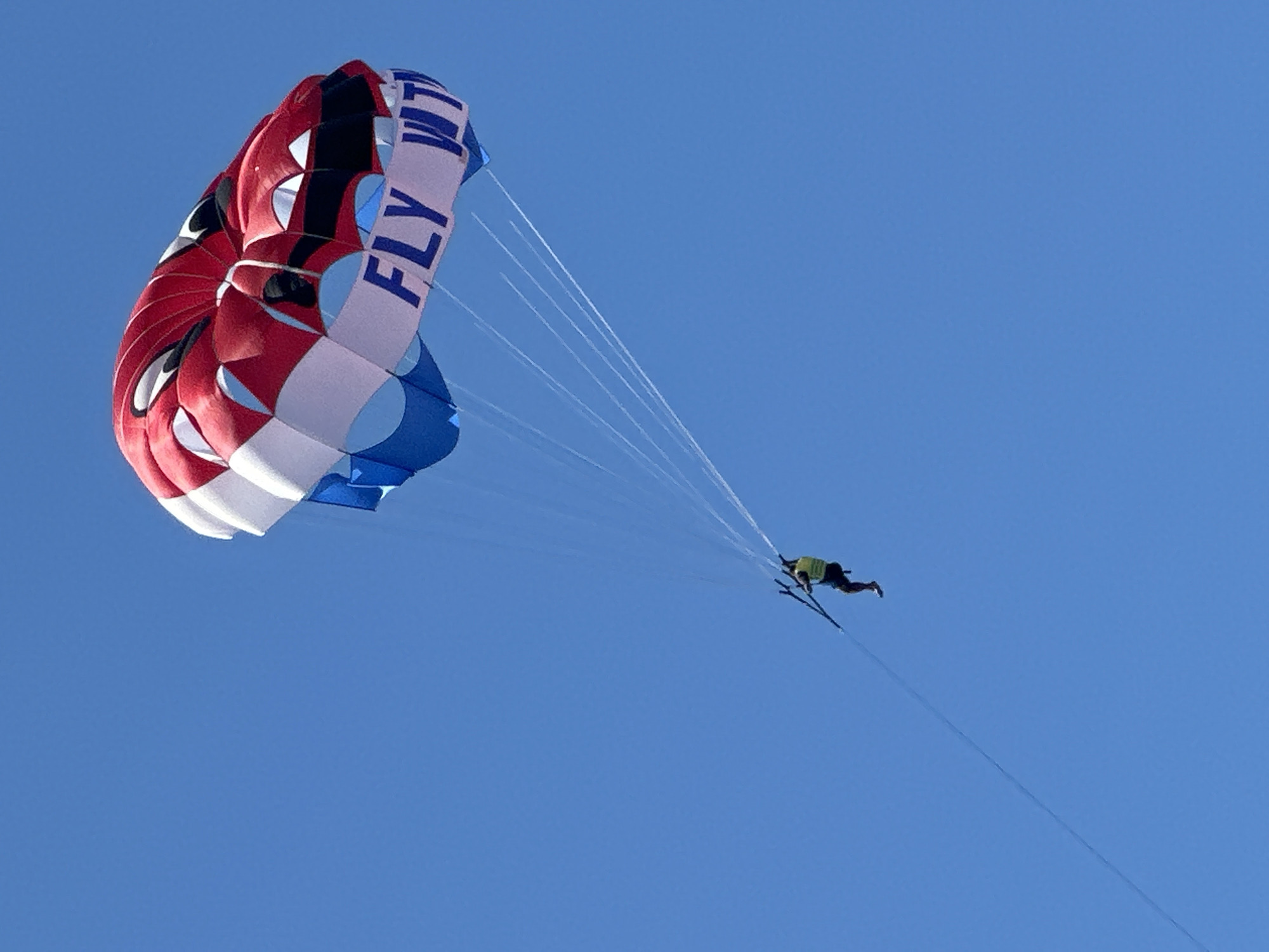 The image captures a parasailer high in a brilliant, cloudless blue sky, with the location context being Budva, Montenegro. The primary subject is a large, colorful parasail, predominantly red, white, and blue, dominating the upper left portion of the frame. Visible on a white section of the parasail is the blue text "FLY WIT" (partially cropped).
Numerous thin lines extend from the parasail, connecting to a single individual suspended in a harness below. This person is wearing a light green or yellow top and dark shorts, and notably appears to have skis or ski-like footwear attached to their feet. A long, taut tow line extends downwards from the individual, out of the frame, indicating they are being pulled by a boat or other vehicle.
The activity depicted is parasailing, with a unique element of the person wearing skis. The bright, clear lighting suggests it is daytime under sunny weather conditions. The photograph is taken from a low-angle perspective, looking upwards at the airborne participant and their colorful parachute against the vast expanse of the sky.