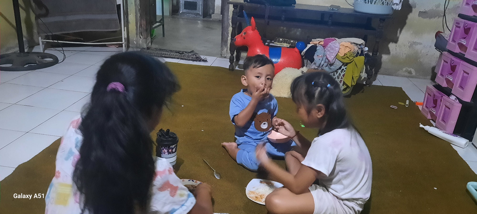 In a room in Ploso, Indonesia, three children are on the floor eating. The child in the center, wearing a blue t-shirt with a teddy bear design, is eating with their hands. To the right, a girl in a white t-shirt is feeding another child, also possibly a girl, who is out of frame. There are plates of food on the floor, including rice. In the background, a red inflatable unicorn toy, laundry in a basket, and a plastic storage unit with pink drawers are visible. The floor is tiled in the foreground and covered with a brown rug in the background.