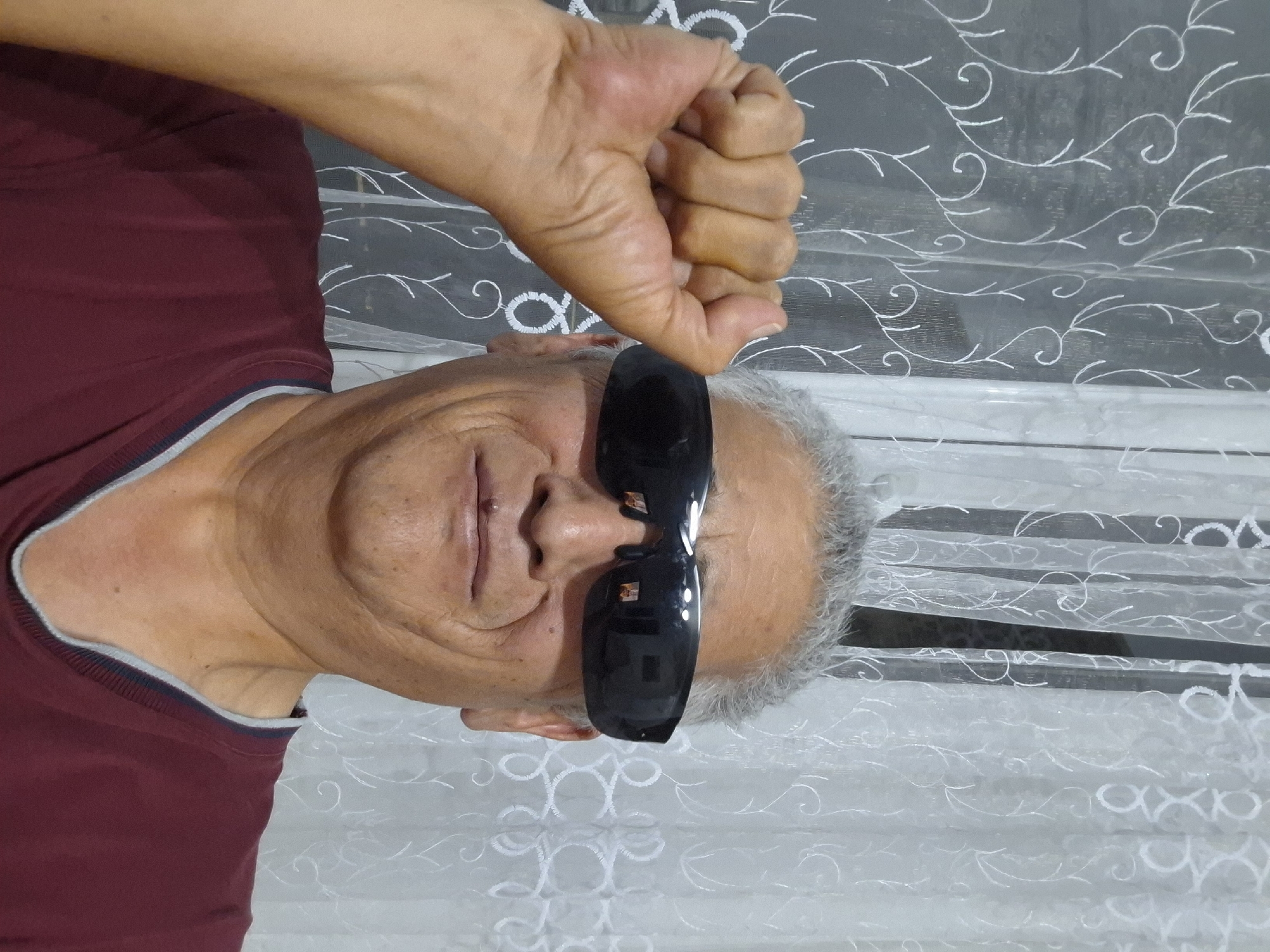 A middle-aged man with graying hair and sunglasses is posing for a photo in front of a white curtain with a floral pattern. He is wearing a maroon shirt with a gray trim around the neckline. His left fist is raised to the side of his face, obscuring part of his head. The lighting suggests an indoor setting, possibly a home. The image was taken in Almaty, Kazakhstan.
