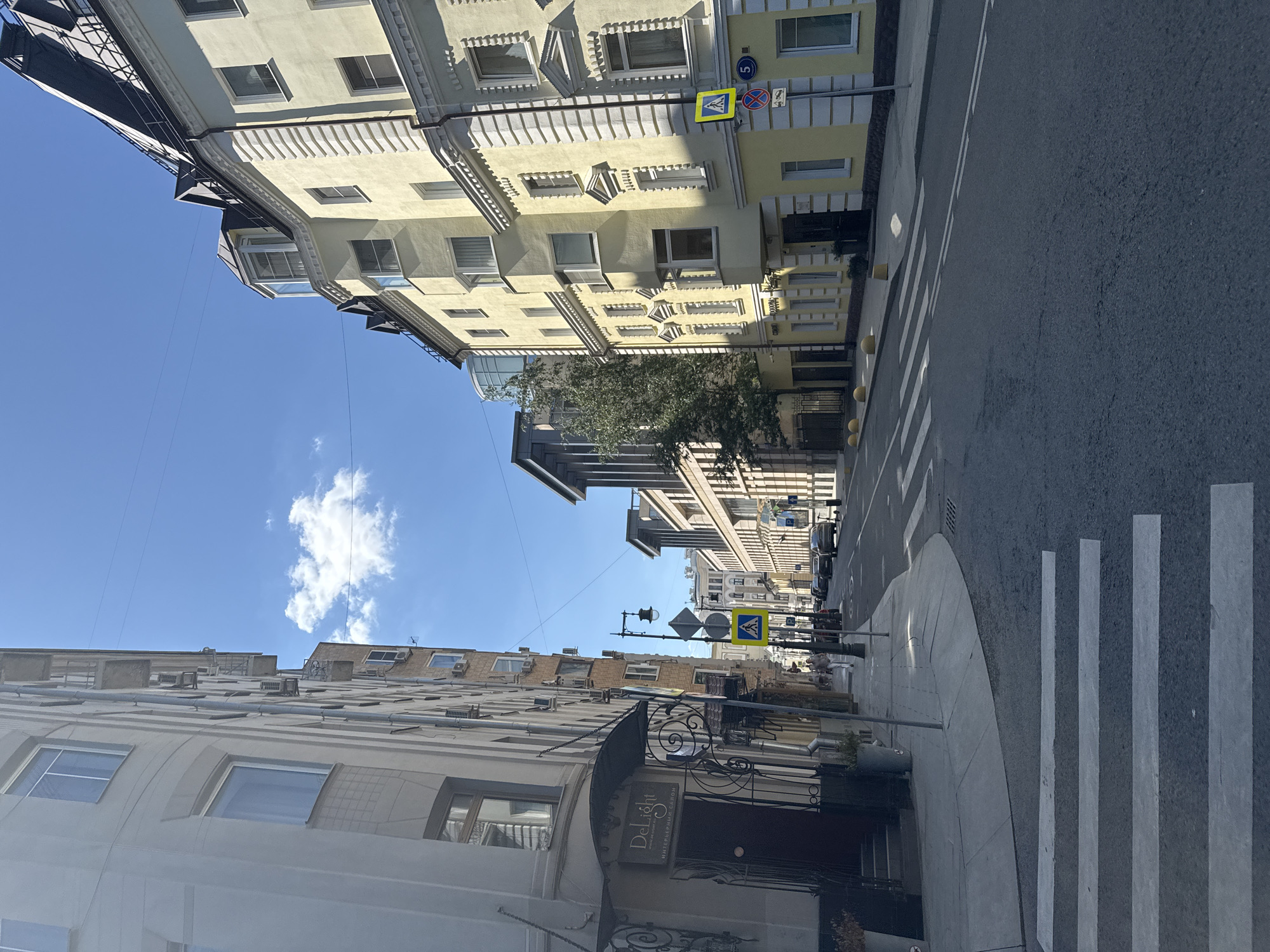 This is a full-frame, eye-level, outdoor, vertical shot of a street scene in Moscow, Russia, on a clear, sunny day. The perspective is from the middle of a road, looking down a street lined with multi-story buildings. On the left is a light-colored building with many windows, some appearing to be air conditioning units attached. A dark entrance with a metal awning has a sign that reads "Delight" in English and "Интерьерный САЛОН" in Russian. The sidewalk is visible in front of this building.
On the right side of the street, a pale yellow building with white decorative trim and numerous windows dominates the view. It features a rounded bay window and a dark mansard roofline with several dormer windows. Further down the street, more buildings are visible, including some with modern architectural elements.
The street itself is asphalt, marked with double yellow lines in the center and a pedestrian crossing with white stripes in the foreground. Traffic signs are present, including a pedestrian crossing sign with a yellow diamond shape, a circular sign with a blue background and a white number "5", and a circular red and blue sign. Several cars are parked along the right side of the street and a few are driving in the distance. A street lamp with a sign attached is also visible.
A single person, a woman, is walking on the sidewalk on the right side of the street, moving away from the camera. The sky is a bright blue with a single white cloud. The overall impression is a typical urban street on a pleasant day.