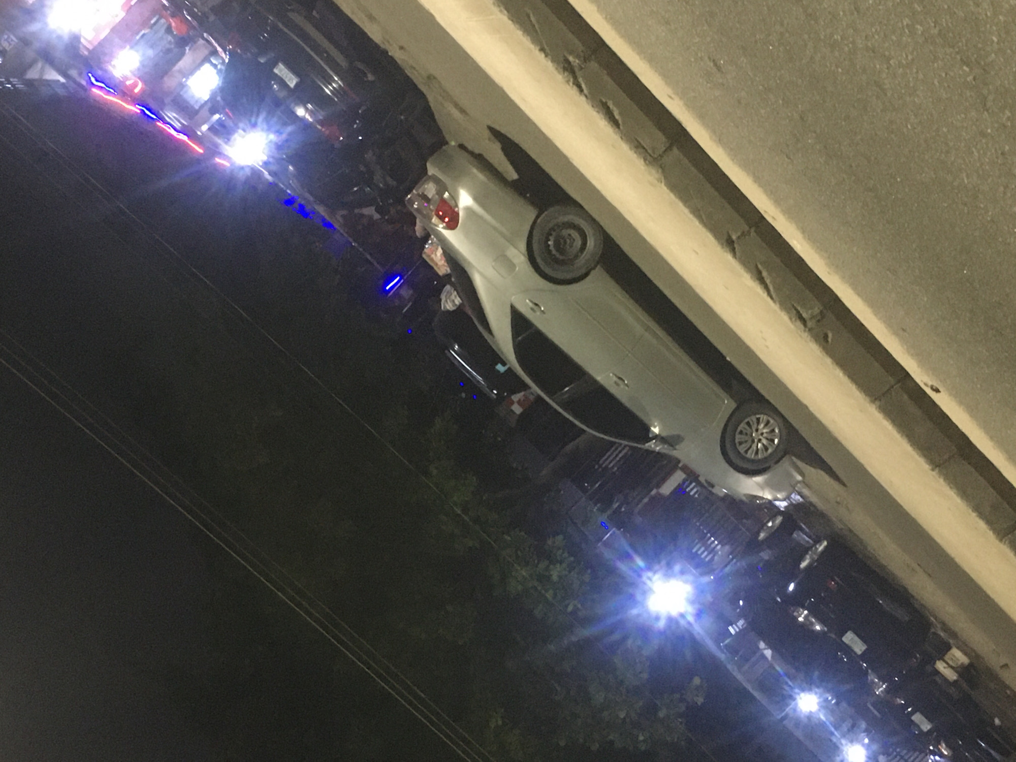 At night in Ado, Nigeria, a silver car parks along a curb. Several dark vehicles are parked nearby, with lit headlights. A few people are visible around the vehicles. Power lines run across the night sky. Vegetation is visible near the vehicles.