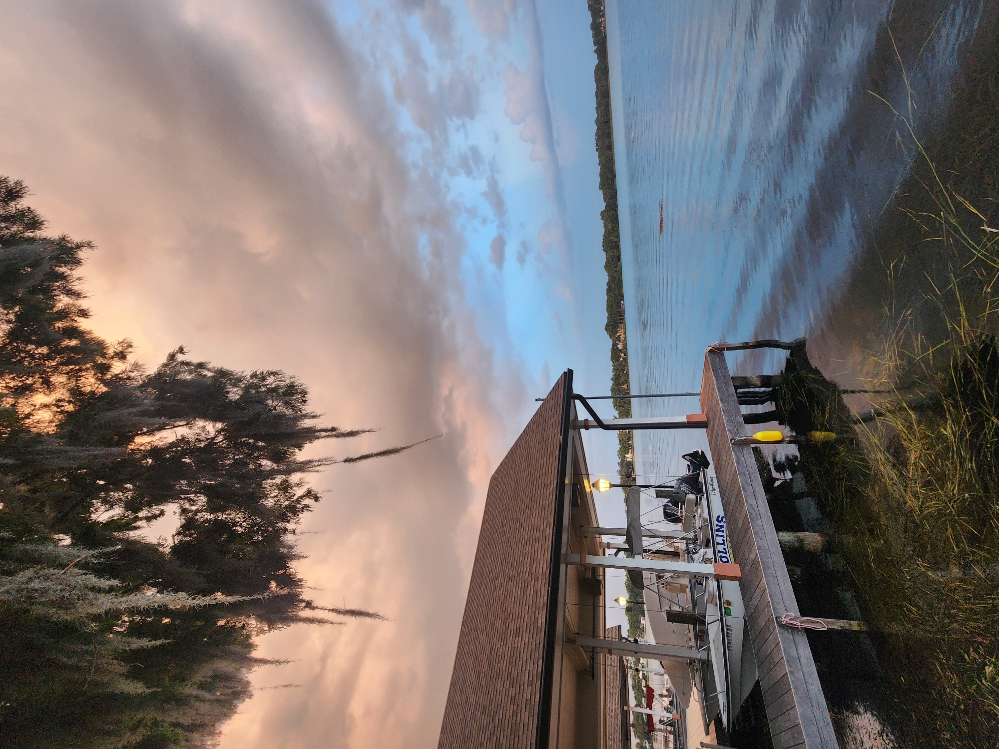 This image captures a serene sunset scene at a boat dock on a lake in Winter Park, Florida. In the foreground, a wooden dock extends into the calm water, where aquatic plants are visible beneath the surface. Two boats are moored under a covered dock. Spanish moss hangs from a tree branch in the upper left corner, silhouetted against a sky painted with hues of pink, orange, and blue. The water reflects the colorful sky, creating a peaceful and tranquil atmosphere. The background features a distant treeline and houses, suggesting a residential area along the lake.