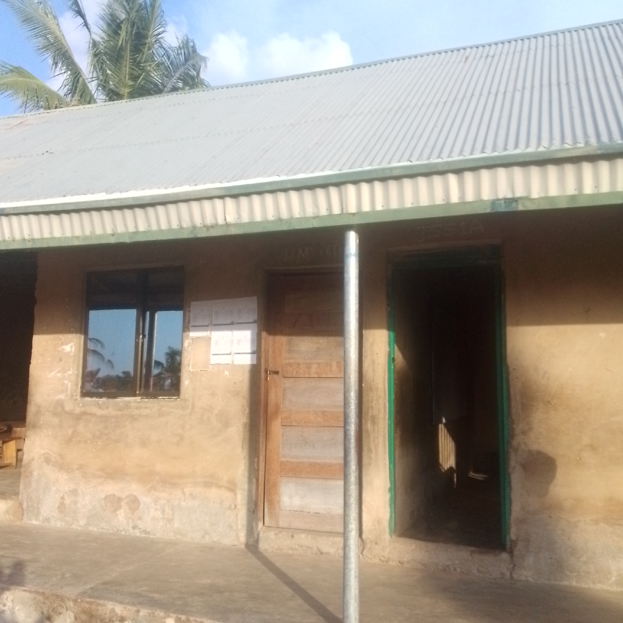 The image shows a simple, single-story building with a corrugated metal roof. The walls are made of a tan-colored material, possibly stucco or plaster, and appear somewhat worn. To the left, there is a window with dark-tinted glass, reflecting palm trees and a blue sky with white clouds. To the right of the window, several pieces of white paper are affixed to the wall.
In the center of the building, there is a wooden door with horizontal panels, and next to it, a vertical metal pole. To the right of the pole, there is an open doorway with a green frame, leading into a dark interior. Above this doorway, the text "JS51A" is visible in white letters. The scene suggests a casual, perhaps educational or administrative, setting. The bright sky and shadows indicate it is daytime, likely sunny. The presence of palm trees suggests a tropical or subtropical location. The overall impression is one of a functional, unpretentious structure.