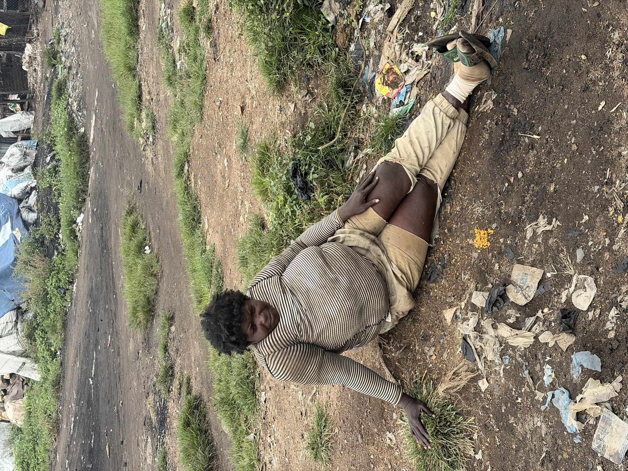 A person is sitting on the ground in what appears to be a disposal site in Bukuru, Nigeria. The individual has dark skin, short dark curly hair, and is wearing a striped long-sleeved shirt and tan shorts. The person is leaning on their left arm with their legs extended. The ground is covered in dirt, debris, and trash, including plastic bags, paper, and other refuse. In the background, there are some structures and more piles of garbage. The overall environment appears unkempt and is characteristic of a waste dump or informal settlement.