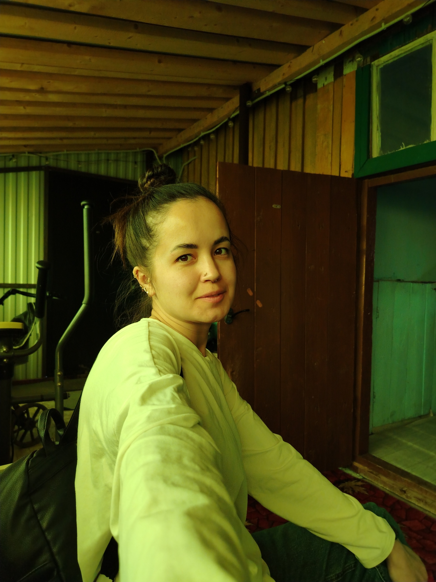 A medium shot captures a woman inside what appears to be a rustic, possibly rural, structure. The woman has her dark hair pulled back in a bun and is wearing a light-colored long-sleeved shirt. She is looking towards the camera with a slight smile. She has multiple piercings in her ear and a nose ring.
The background reveals a space with wooden walls and ceiling. The lighting is dim and casts a yellow tone over the scene. To the right is a partially open brown wooden door, revealing a green-toned interior. Behind the woman, an exercise machine can be seen. A dark backpack is draped on the chair.
The location appears to be Гожан, Russia, based on the user's prompt. The environment suggests a simple, possibly rural or cottage-like dwelling.