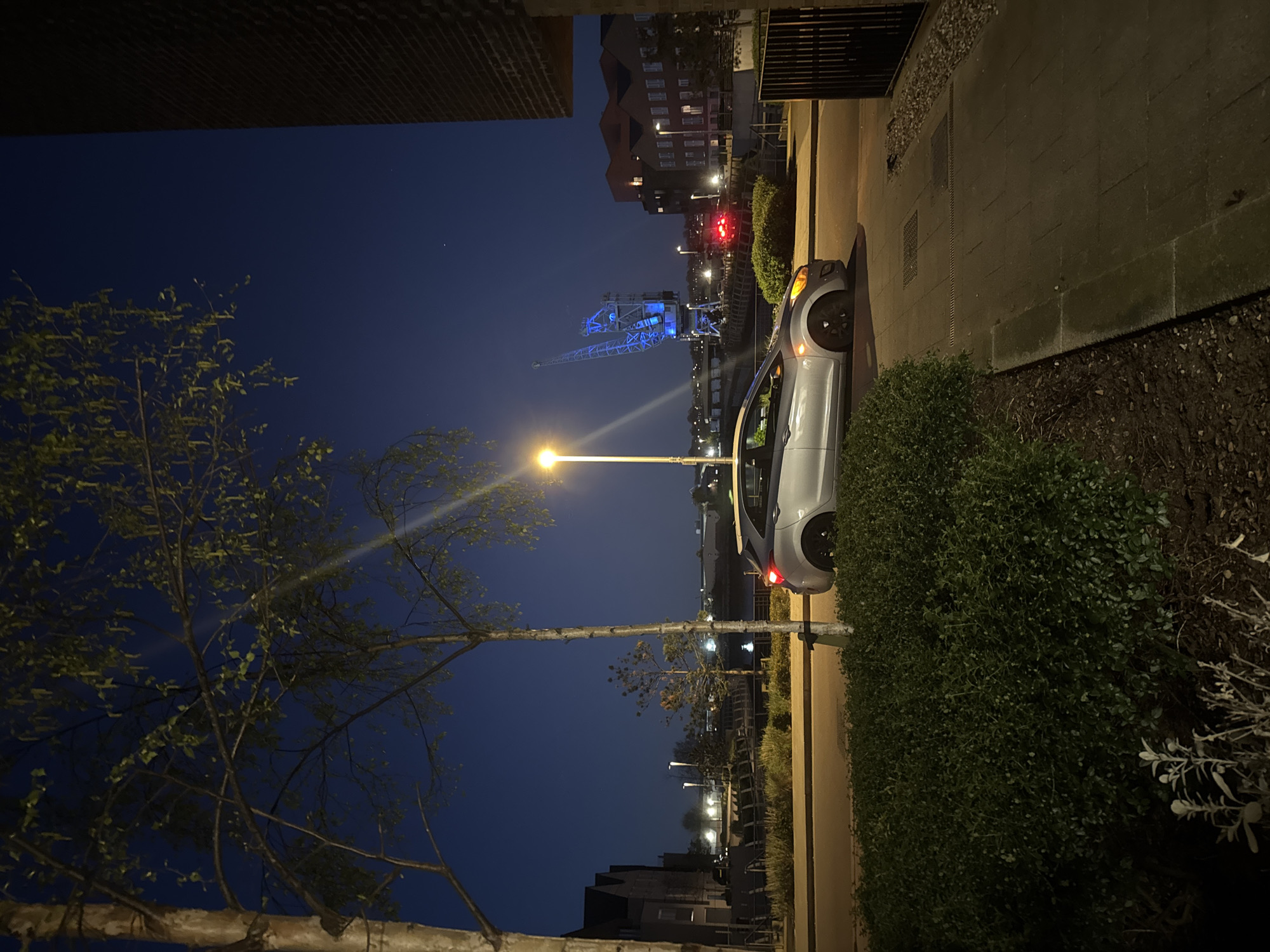 The image depicts a nighttime scene in Rochester, United Kingdom. A silver car is parked on a paved street next to a building and a substantial hedge. In the distance, a brightly lit blue crane, a local landmark, is visible against the dark sky. Streetlights illuminate the scene, casting light on the buildings and foliage. A tree with sparse leaves is on the left side of the image. The sky is a deep blue, suggesting it is late evening or night. The location appears to be a residential area within the city, close to the riverfront.