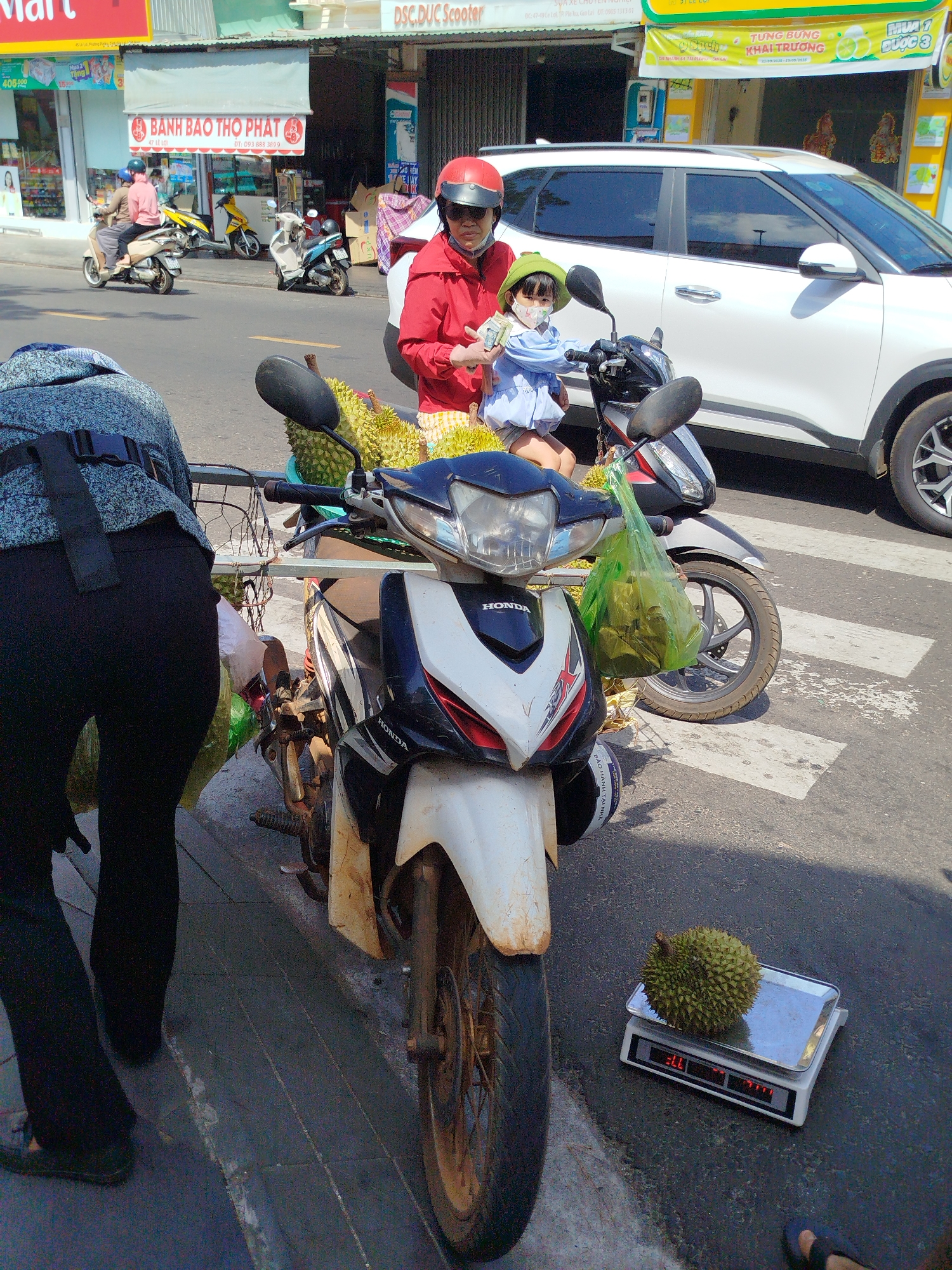 A woman and a child, both wearing helmets, sit on a scooter parked on the side of the road in Pleiku Ward, Vietnam. Durian fruits are loaded onto the scooter. A person in black pants leans over, possibly making a purchase. One durian sits on a digital scale on the ground. In the background, there is traffic and storefronts, including "Banh Bao Tho Phat" and "DSC Duc Scooter." A white SUV is parked nearby.