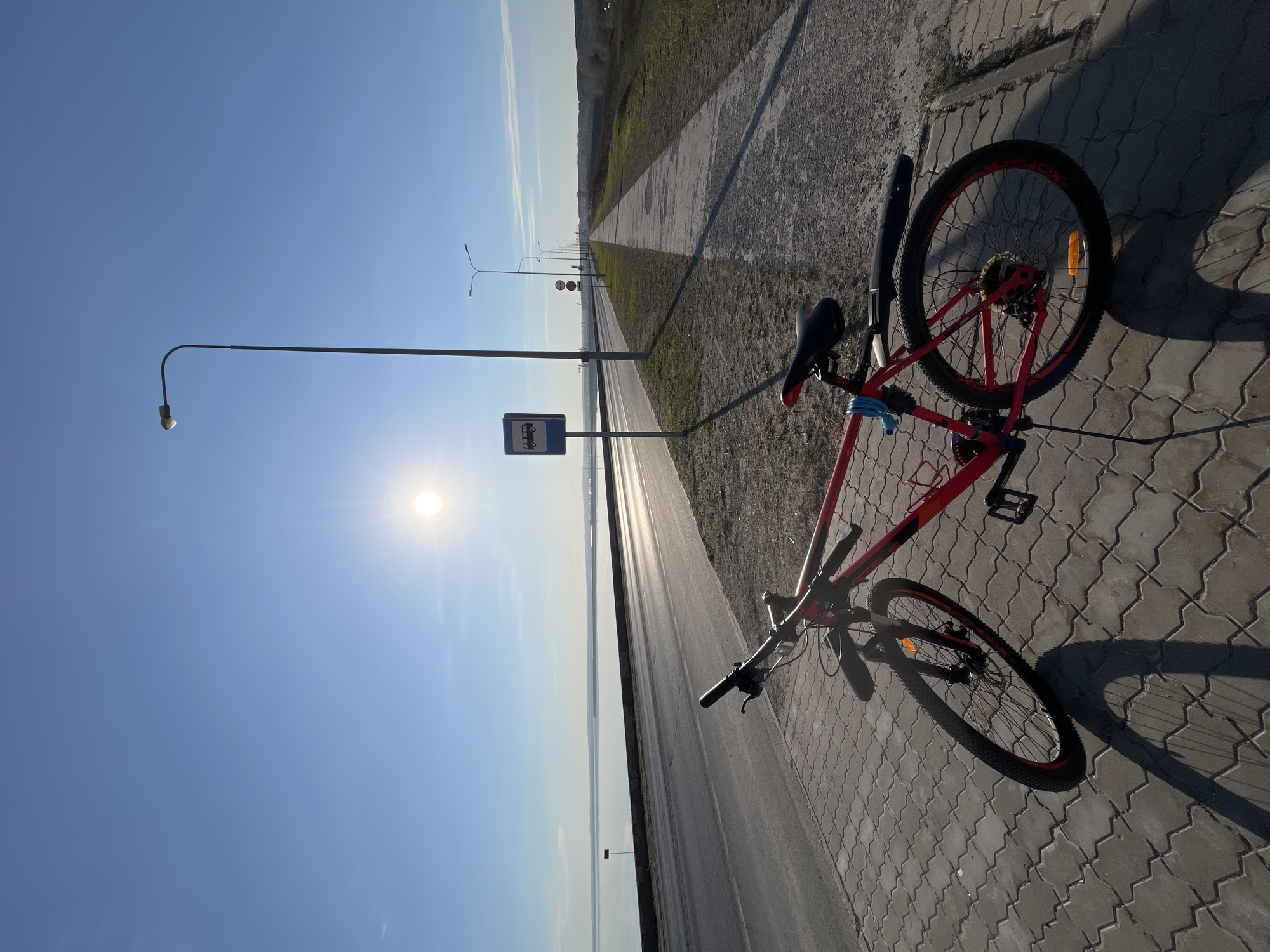 The image shows a red mountain bicycle lying on its side on a paved sidewalk. A blue bus stop sign stands next to the sidewalk, illuminated by the sun. The location appears to be in Chaikovsky, Russia, indicated by the bus stop sign design, the road leading in the distance, and the landscape. Beyond the road and sidewalk, there is a body of water reflecting the sunlight. The sky is clear and blue with the sun brightly positioned. The long shadows suggest it is either early morning or late afternoon. No people are visible.