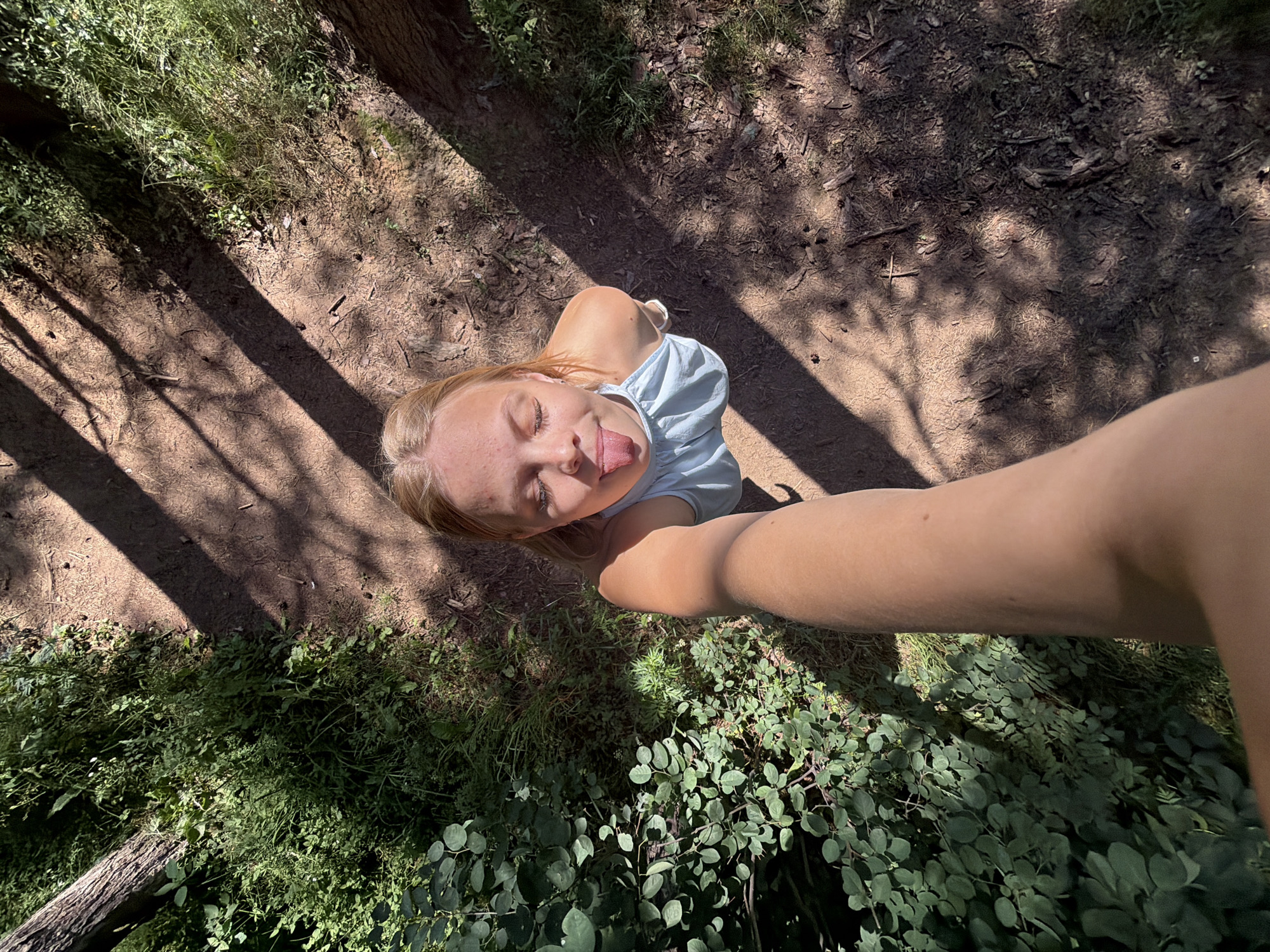 A high-angle, wide shot shows a young woman sticking her tongue out and closing her eyes, as if playing a trick. She is wearing a light blue t-shirt and her arm is extended towards the camera, obscuring part of her face. The scene appears to be outdoors, with dappled sunlight casting shadows on a dirt path. Lush green foliage and trees surround the path, suggesting a wooded or park-like setting. The overall mood is playful and carefree. The image was likely taken during the day, given the sunlight and shadows.