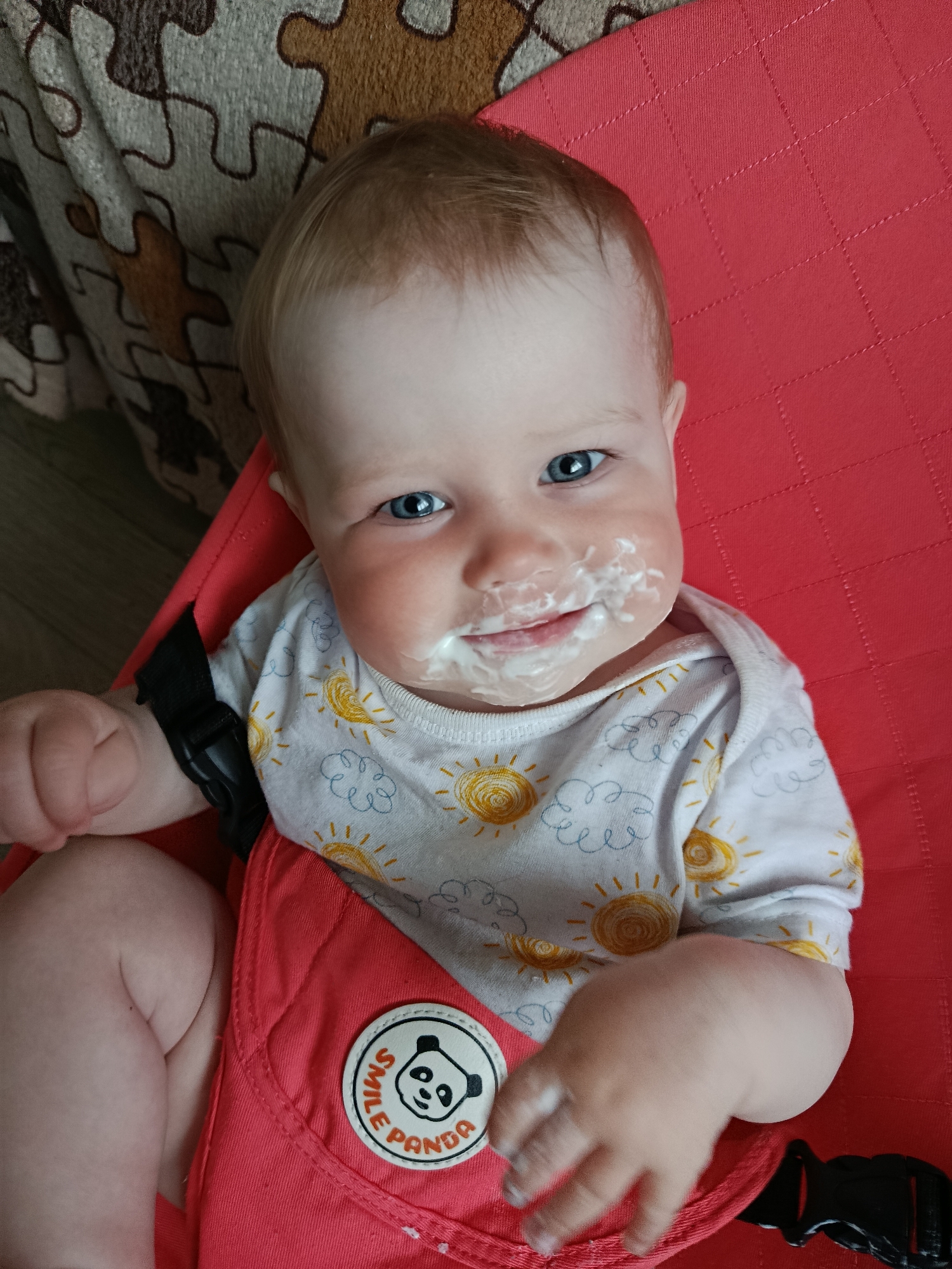 A baby sits in a red baby seat. The baby has light hair and blue eyes, and white food covers the area around their mouth. The baby wears a white shirt with yellow suns and blue clouds printed on it. A patch with a panda and the words "Smile Panda" is on the baby seat. A tan and brown patterned blanket is visible in the upper-left corner. The photo was taken indoors. The location, Фоки, Russia, cannot be determined from the image.