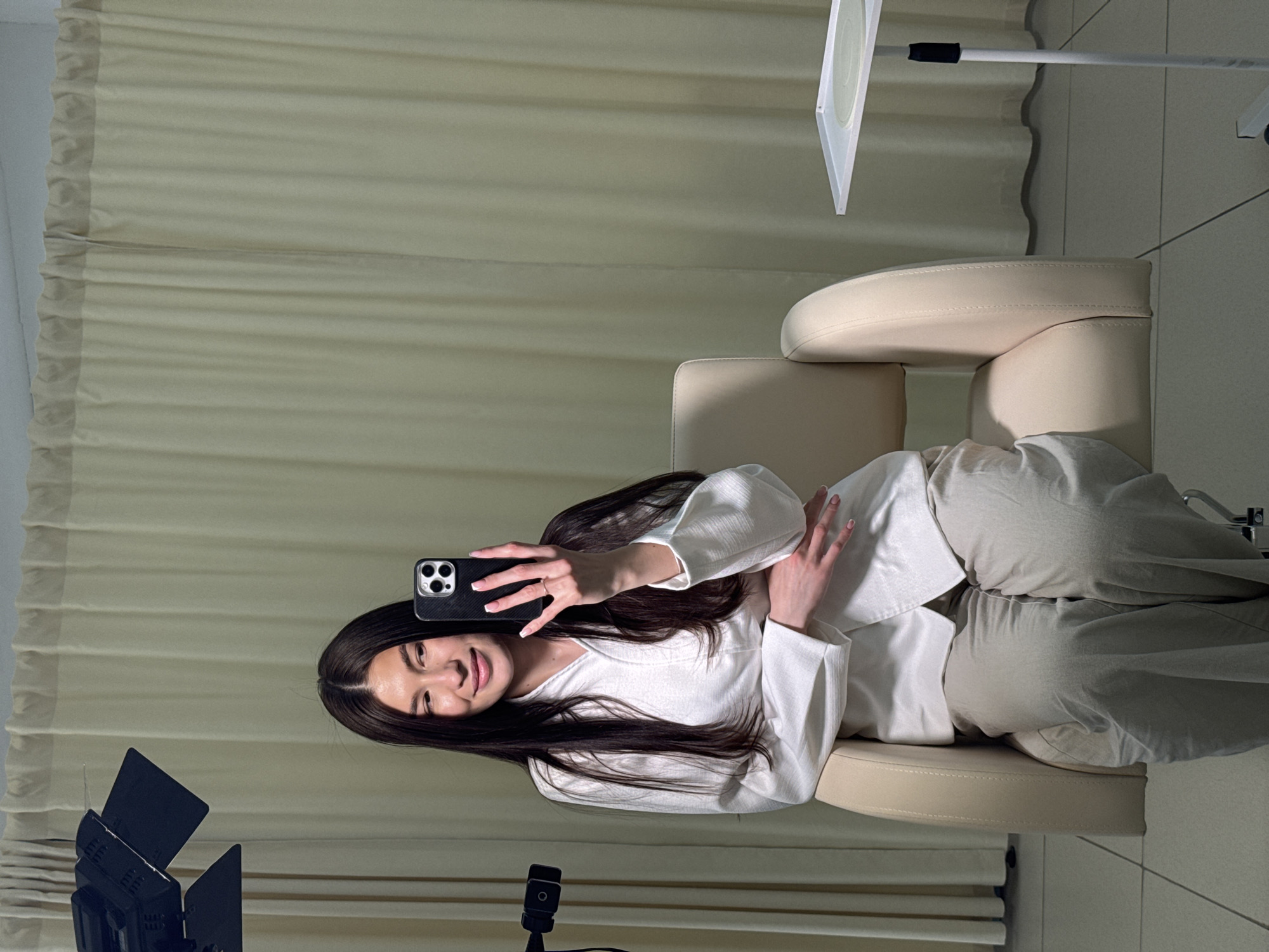 A young woman with long dark hair is seated in a light beige, possibly leather, armchair in an indoor setting. She is holding a smartphone in her right hand, appearing to take a selfie or record a video, and is smiling gently at the camera. Her left hand rests on her waist. She is dressed in a white long-sleeved top and light-colored trousers.
The background features light beige or off-white pleated curtains. To the left of the woman, a black LED panel light on a stand is visible, indicating a controlled lighting setup. To her right, a white stand with a circular reflector or diffuser is also present. The floor appears to be light-toned tile. The overall ambiance suggests a studio environment or a dedicated space for content creation, located in Chaikovsky, Russia. The woman's expression is pleasant and relaxed. There is no visible text in the image.