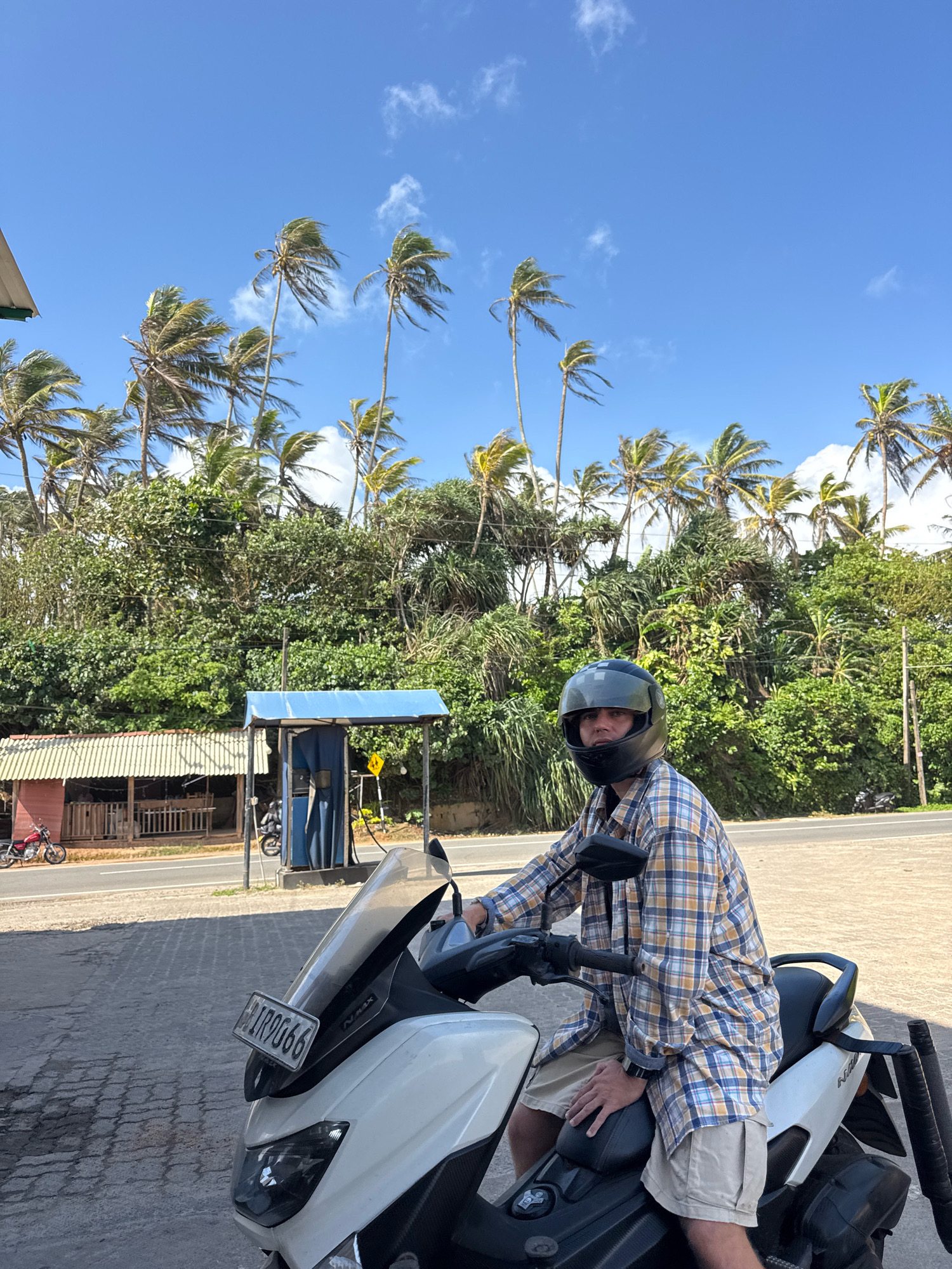 In Goviyapana, Sri Lanka, a person wearing a helmet and a plaid shirt sits on a white scooter. The scooter has a license plate with the number "LIR 90666". Behind the scooter, palm trees and lush green vegetation form a dense backdrop under a bright blue sky with scattered clouds. To the left, a small red motorcycle is parked next to a building with a white roof and wooden structures. A blue-canopied structure, possibly a gas pump, is also visible. The scene suggests a roadside location in a tropical environment.