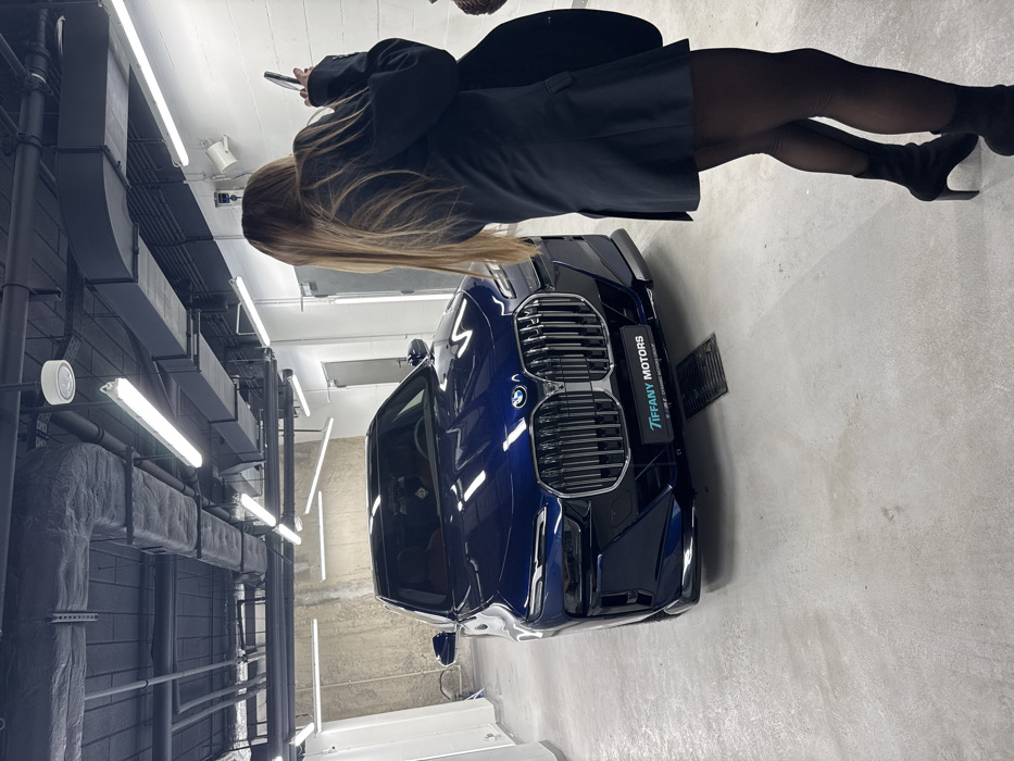 A dark blue BMW automobile, gleaming under fluorescent lights, is prominently displayed. A woman stands beside the car, wearing black tights, heeled boots, and a long black coat. She appears to be looking at her mobile phone. The environment features industrial-style elements, including exposed pipes and a metallic ventilation system. The "Tiffany Motors" logo is visible on the car.