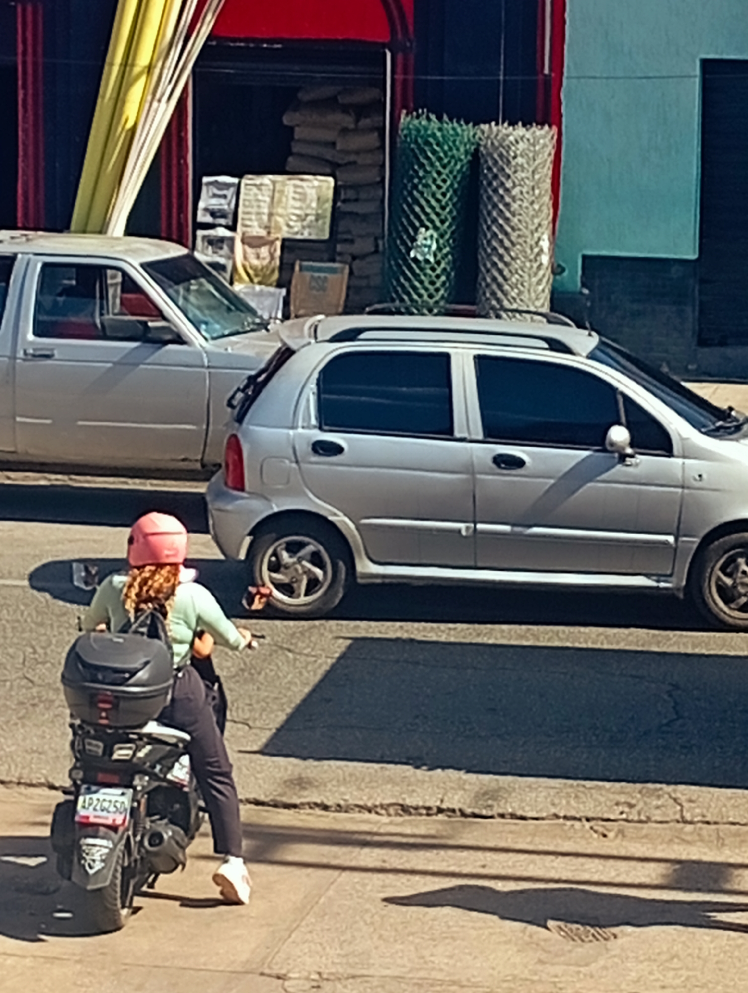 A person wearing a light green long-sleeved shirt, dark pants, white sneakers, and a pink helmet rides a black scooter with a black storage box mounted on the back. The scooter has a license plate that reads "AP2G250". The scooter is positioned on a paved surface in front of a light gray hatchback car, which is parked on a street. Behind the hatchback, a white pickup truck is parked. The background shows a building with a red awning and yellow and red striped curtains. Piled bags of what appear to be cement are stacked next to the building, along with two rolls of green and beige mesh material. The scene is lit by sunlight, casting shadows on the ground. The location is identified as Juan Griego, Venezuela.