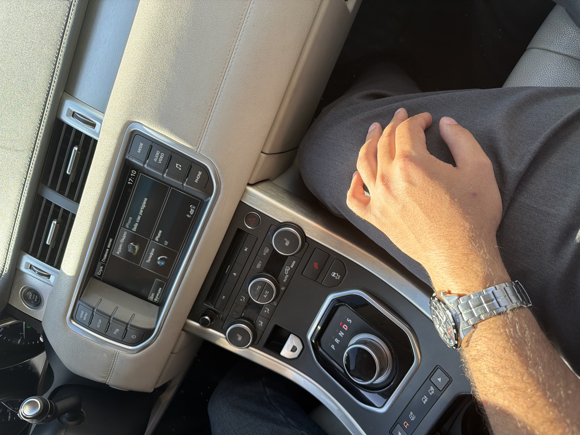 The image captures a high-angle view of a car's interior, focusing on the center console and the upper leg and arm of a person. The scene is set in Krasnoyarsk, Russia, under bright, sunny conditions, indicated by strong highlights and shadows.
A person is seated in the car, wearing grey trousers. Their right hand rests casually on their upper leg, while their left forearm, adorned with a metallic, silver-toned wristwatch, is visible near the gear selector. The watch features a complex, possibly skeletonized dial.
The car's interior features a light beige or tan dashboard and upper console made of a soft-touch material, possibly leather, with subtle stitching. Brushed metallic accents are integrated into the design, particularly around the infotainment screen and climate control panel. The infotainment screen displays "17:10" (the time), and various options in Russian, including "Служ. Главное меню" (Service. Main menu), "Аудио/Видео" (Audio/Video), "Телефон" (Phone), and "Доп. функции" (Additional functions). The screen also shows "iPhone" twice and "Bella ciao partigiana," likely a song title currently playing or selected. Buttons around the screen include "MODE," "AUDIO VIDEO," and "PHONE."
Below the infotainment screen are physical controls for the climate system, featuring circular dials and buttons for "AUTO," "A/C," and heated seats, among others. A rotary gear selector is prominent, displaying "P R N D S" (Park, Reverse, Neutral, Drive, Sport), currently in the "P" (Park) position. Other visible controls include a "START STOP" button, "HOME MENU," "SETUP," and hazard lights. Air vents are visible on the dashboard to the left.
