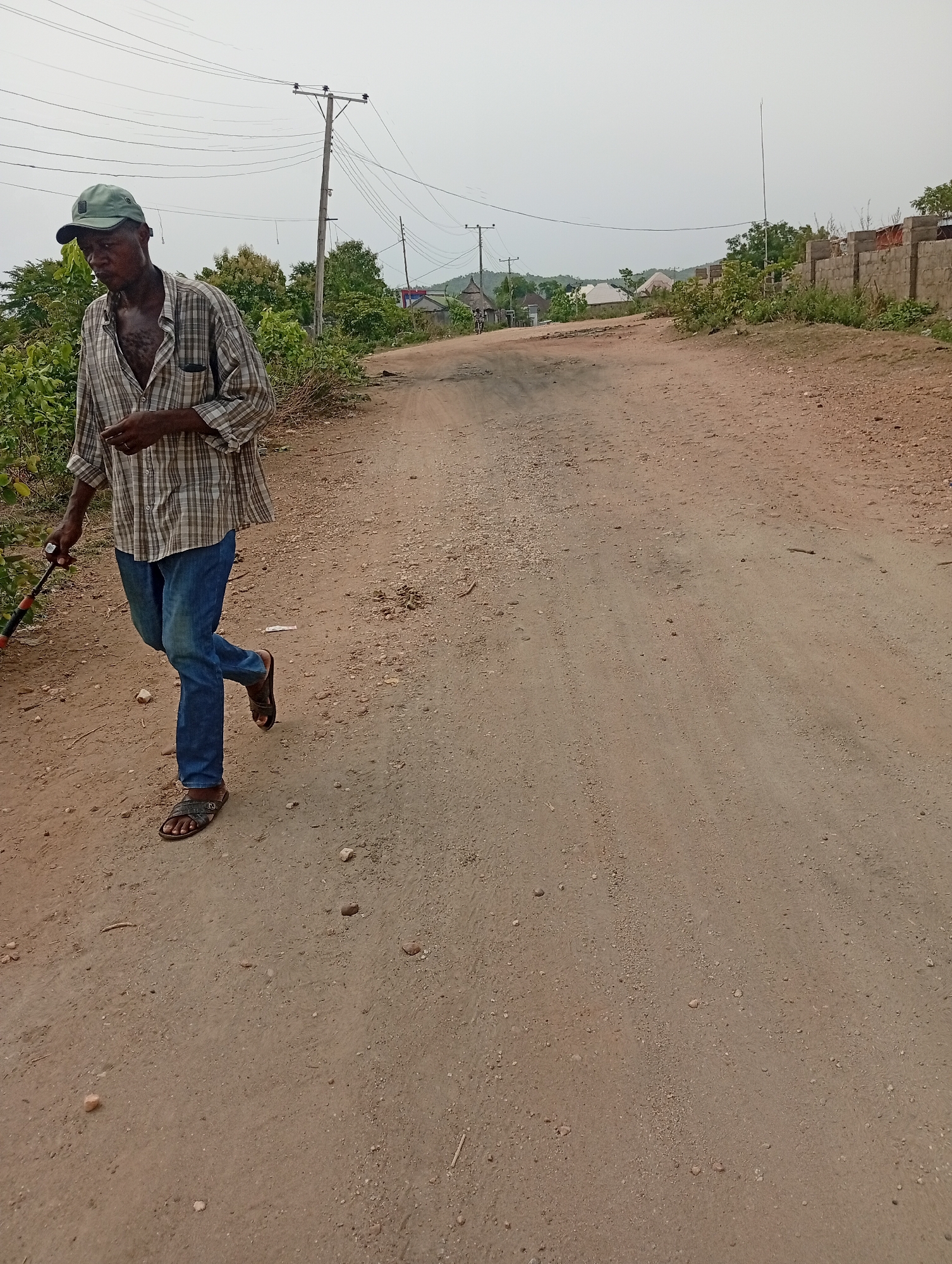 A man walks along a dirt road in Adankolo, Nigeria. The man wears a plaid shirt, blue jeans, sandals, and a green cap. He carries a stick. The road is unpaved, with vegetation on either side. Utility poles line the road. In the background are buildings and hills. The sky is overcast. A partially constructed wall is visible on the right side of the road.