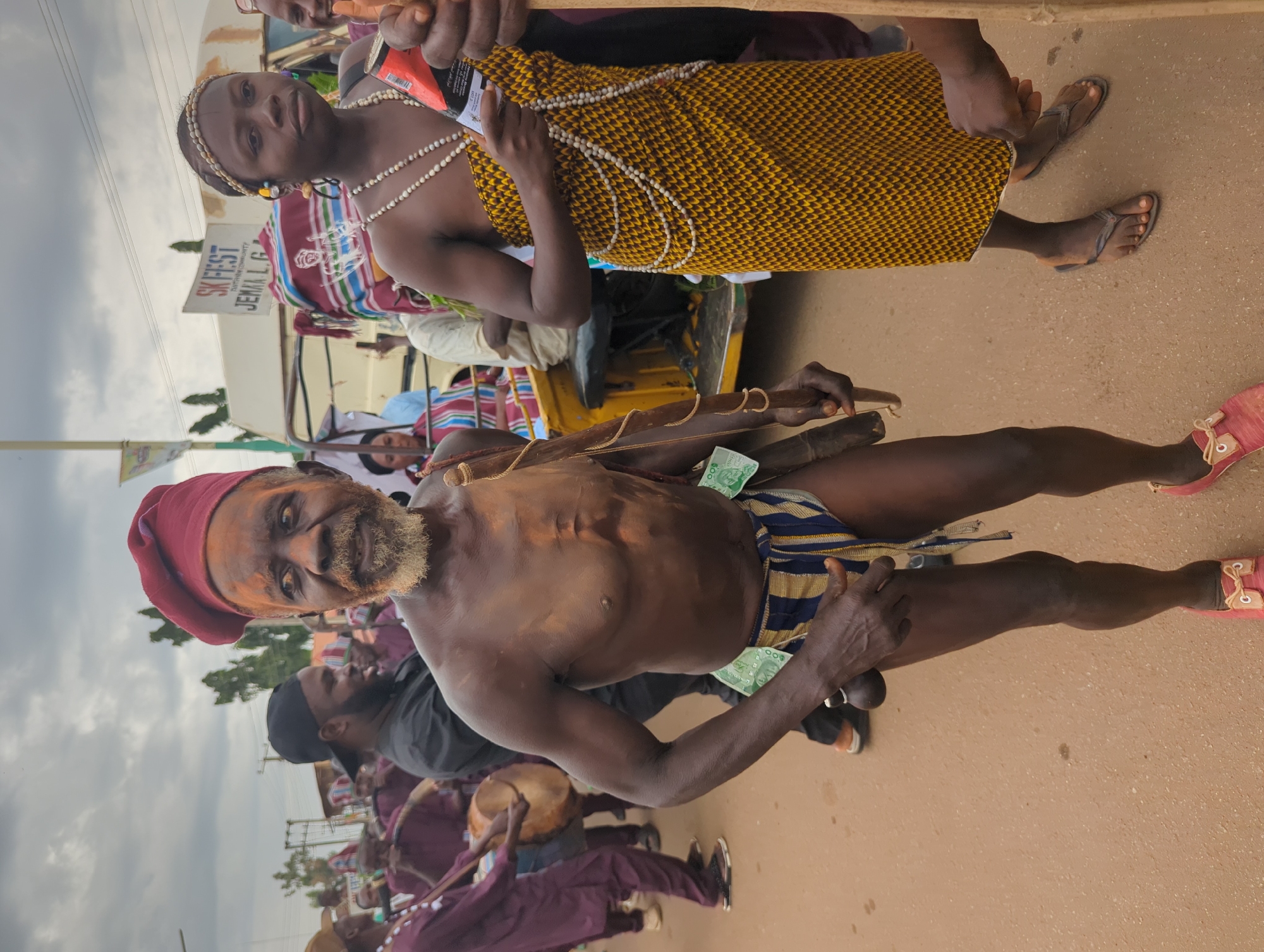 The image depicts a scene from Maigizo, Nigeria, featuring two main subjects and a group of musicians. The foreground shows a shirtless man wearing a red cap, striped shorts, and red shoes. He has a beard and is holding an instrument made of wood. Money is tucked into his shorts. Behind him, a group of people are playing drums and wearing maroon clothing. Further back, a woman in a yellow patterned dress and white beaded necklaces is visible, sitting in a yellow vehicle. A sign reading "SKIFEST JEMA'A L.G." is partially visible near the vehicle. The background includes a sky with overcast clouds and buildings. The ground is paved.