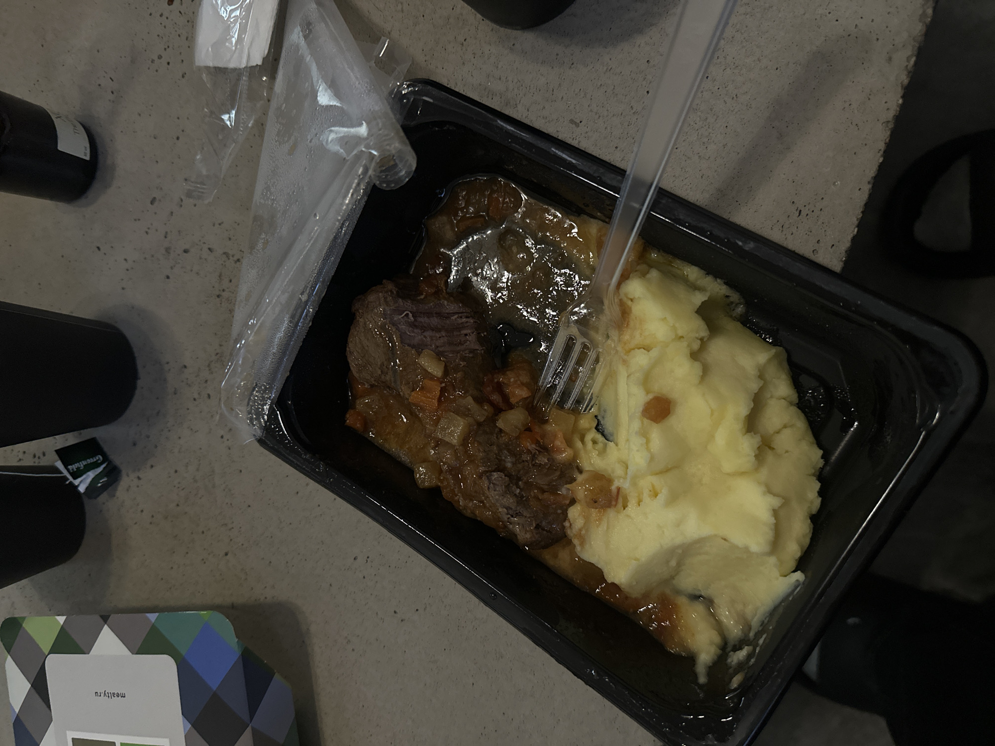 The image shows a ready-to-eat meal in a black plastic container. The meal appears to be beef stew with mashed potatoes. A clear plastic fork is inserted into the meal, partially mixed with the potatoes and stew. The container is partially covered with clear plastic wrap. Beside the container is a small Greenfield tea bag wrapper, a dark-colored bottle (possibly a drink), and a package with a geometric design and the text "maatly.ru". The scene is set on a gray, textured surface, possibly a table or counter. Based on the website maatly.ru, this image could be from Moscow, Russia.