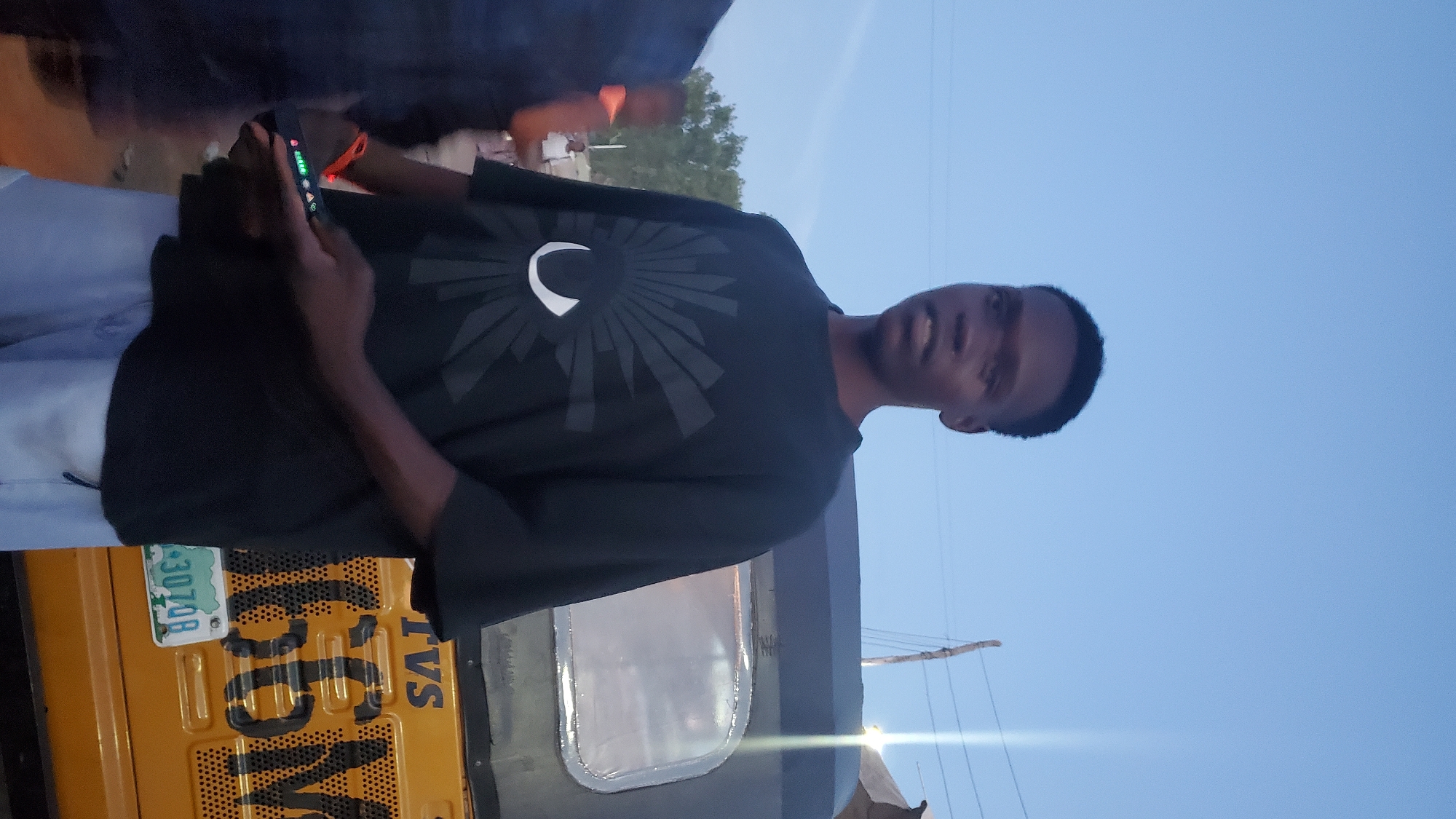 A young Black male stands outdoors, facing forward and slightly to his left, in a dimly lit environment with a clear blue sky overhead. He is wearing a black t-shirt with a stylized white eye graphic and light blue shorts. His left hand holds a black electronic device with a lit screen displaying colorful indicators. Behind him, to his right, is the rear of a yellow three-wheeled vehicle, likely a Bajaj auto-rickshaw, with a license plate visible that reads "NG 3070B". The vehicle has the brand "TVS" and the number "66M" painted on its rear. To the left of the auto-rickshaw, another person is partially visible, blurred and wearing an orange garment. Further in the background, trees are visible, suggesting a semi-urban or rural setting. The overall scene appears to be captured at dusk or dawn.