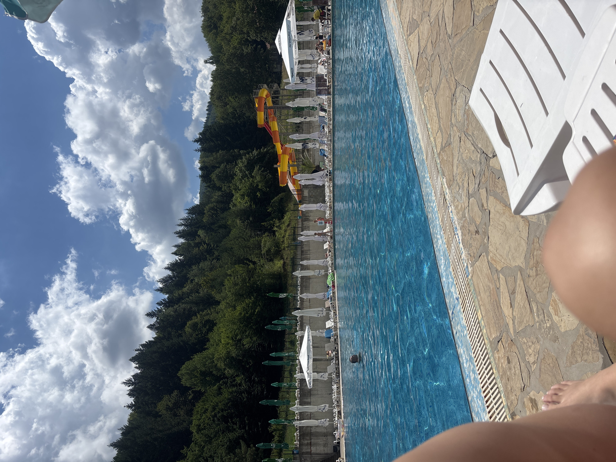 The image depicts an outdoor swimming pool area at a resort in Chiflik, Bulgaria. The pool is large and filled with clear blue water. In the background, a lush green forest covers a hillside under a bright blue sky with fluffy white clouds. To the right of the pool, a colorful water slide with yellow and orange tubes is visible. Several rows of lounge chairs and umbrellas are set up along the pool deck, with some people lounging or walking around. A man is visible in the water, swimming. The foreground shows a portion of a person's legs and feet, suggesting they are sitting on a lounge chair or at the edge of the pool. The overall scene conveys a sense of a relaxing summer day at a recreational facility.
