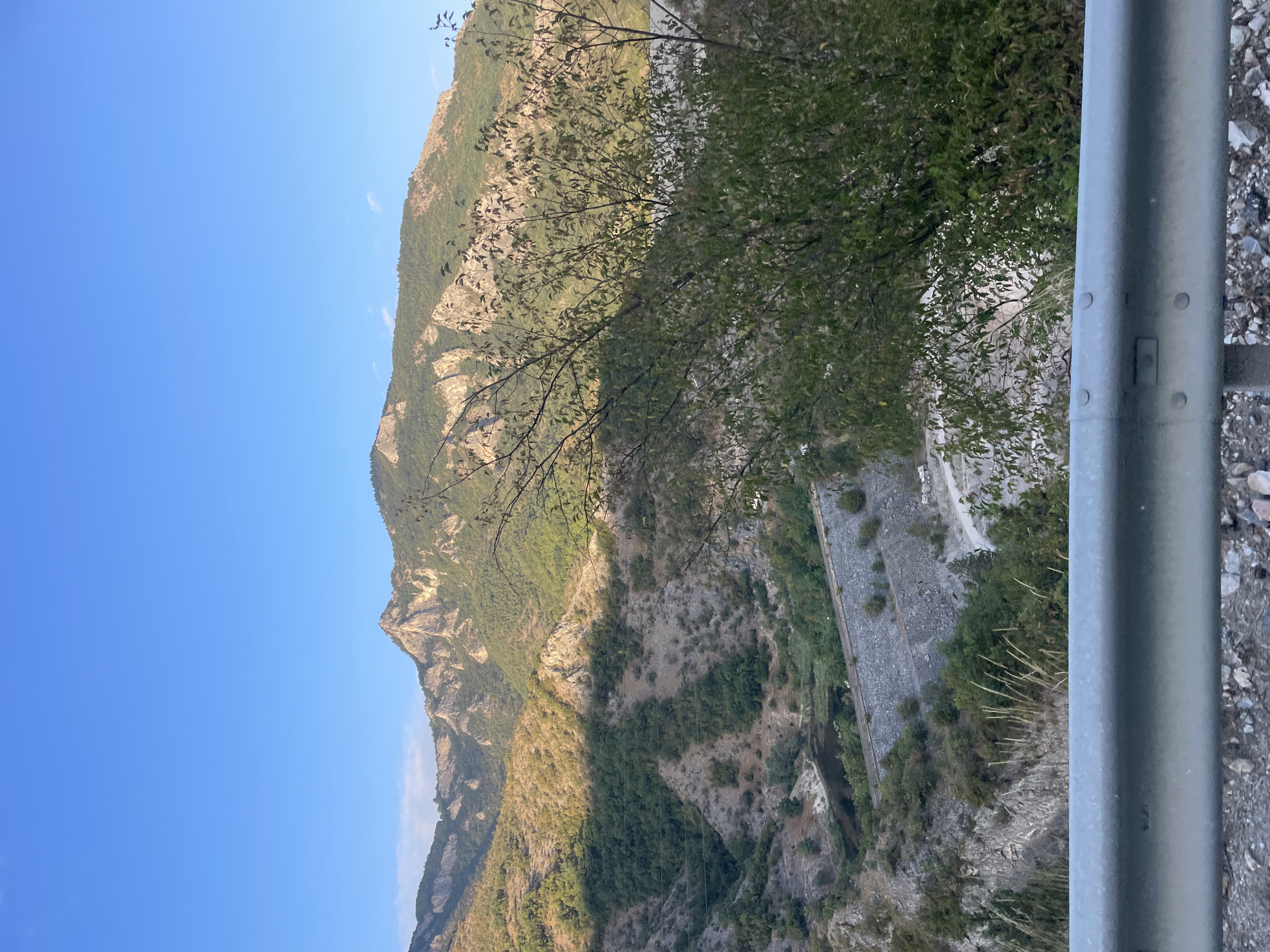 This is a landscape photograph taken from a high vantage point, looking down into a valley with mountains in the background. The sky is a clear, bright blue. The mountains are covered in green trees and rocky outcrops. In the valley, there is a body of dark water, possibly a river or a lake, with a concrete structure, likely a dam, running along its edge. A road is visible on the right side of the dam. In the foreground, there is a metal guardrail, indicating the photo was taken from a road or a viewing platform. The scene depicts a natural environment, possibly a rural or mountainous area, near the city of Yugovo, Bulgaria.