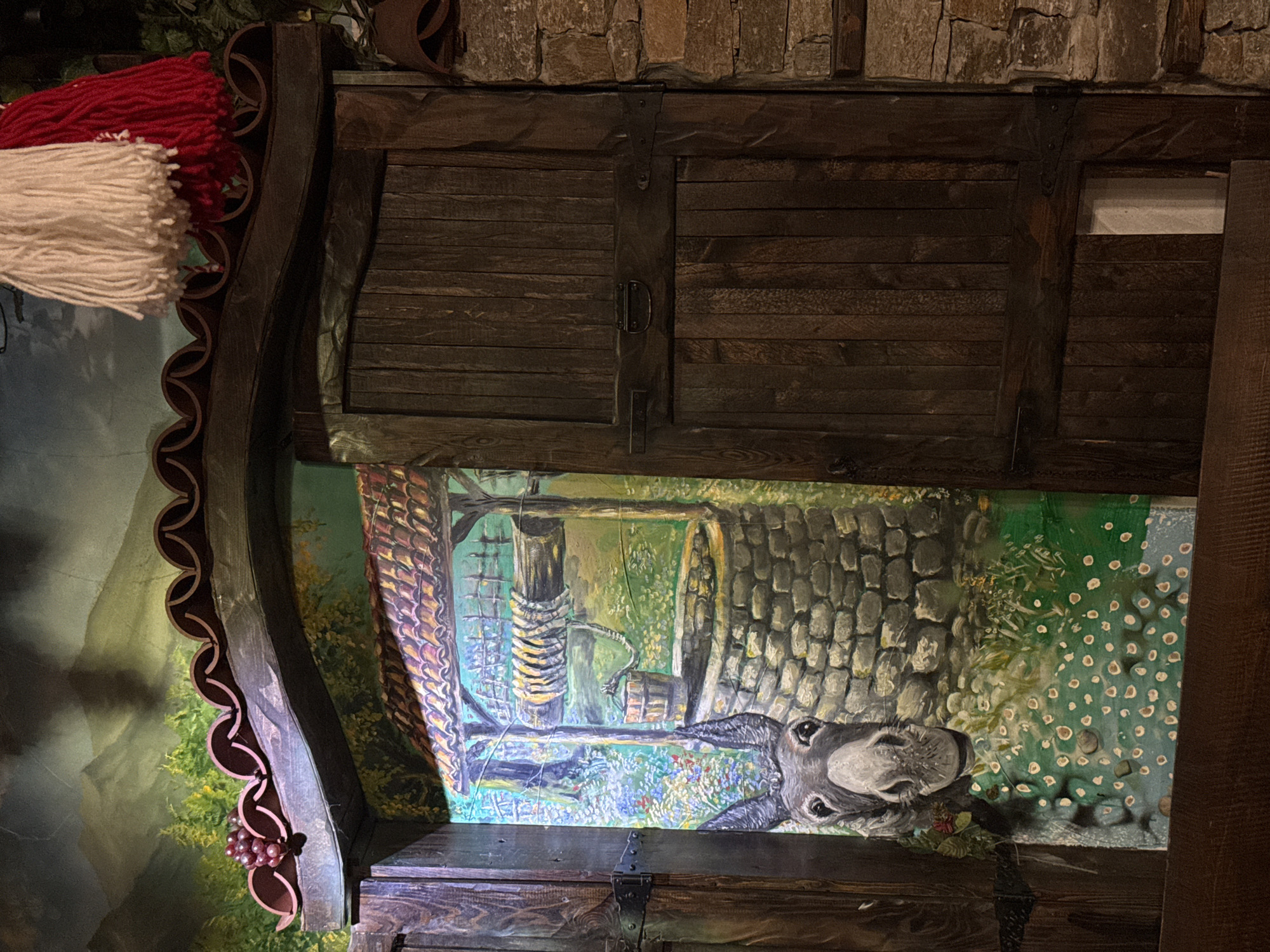 The image depicts a mounted display, possibly within a restaurant. At the top, there is a cabinet made of dark-stained wood with two hinged doors secured by a central clasp. Above the cabinet, a wall made of roughly hewn stones is visible. Below the cabinet is a painting, framed by a dark wood casing with decorative scallops. The painting depicts a scene with a well and a donkey; the perspective is as if the viewer is looking down into the well. Adjacent to the frame, on the upper left, is a decorative fringe of white and red yarn. The overall aesthetic suggests a rustic, European-themed establishment.
Given the specific request for a location in Des Plaines, United States of America, and knowing the decor style, it may be located in a restaurant or tavern there.