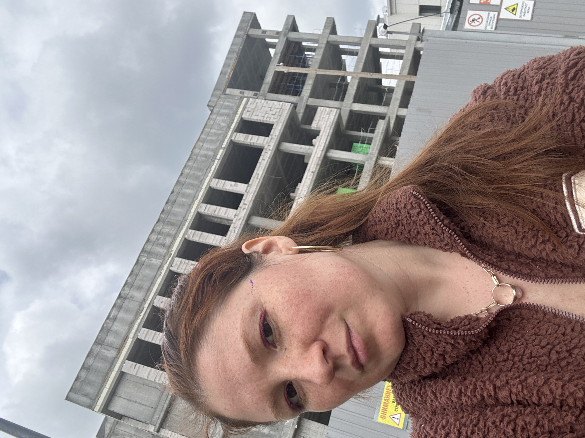 A medium shot depicts a woman with light skin, freckles, and reddish-brown hair, positioned in front of a concrete structure under construction. She is wearing a brown, textured, zip-up jacket with a decorative chain accent at the zipper. Her makeup includes pink eyeliner and what appears to be a small, faded tattoo or marking on her forehead near her left eyebrow. She is wearing large hoop earrings.
Behind her is an unfinished building with a visible concrete framework. A grey sky with clouds is visible above the structure. To the right of the building, a grey, metallic fence or barrier is present with a yellow warning sign attached. The sign contains Russian text. Based on contextual clues, the location is presumed to be in Nizhny Novgorod, Russia.