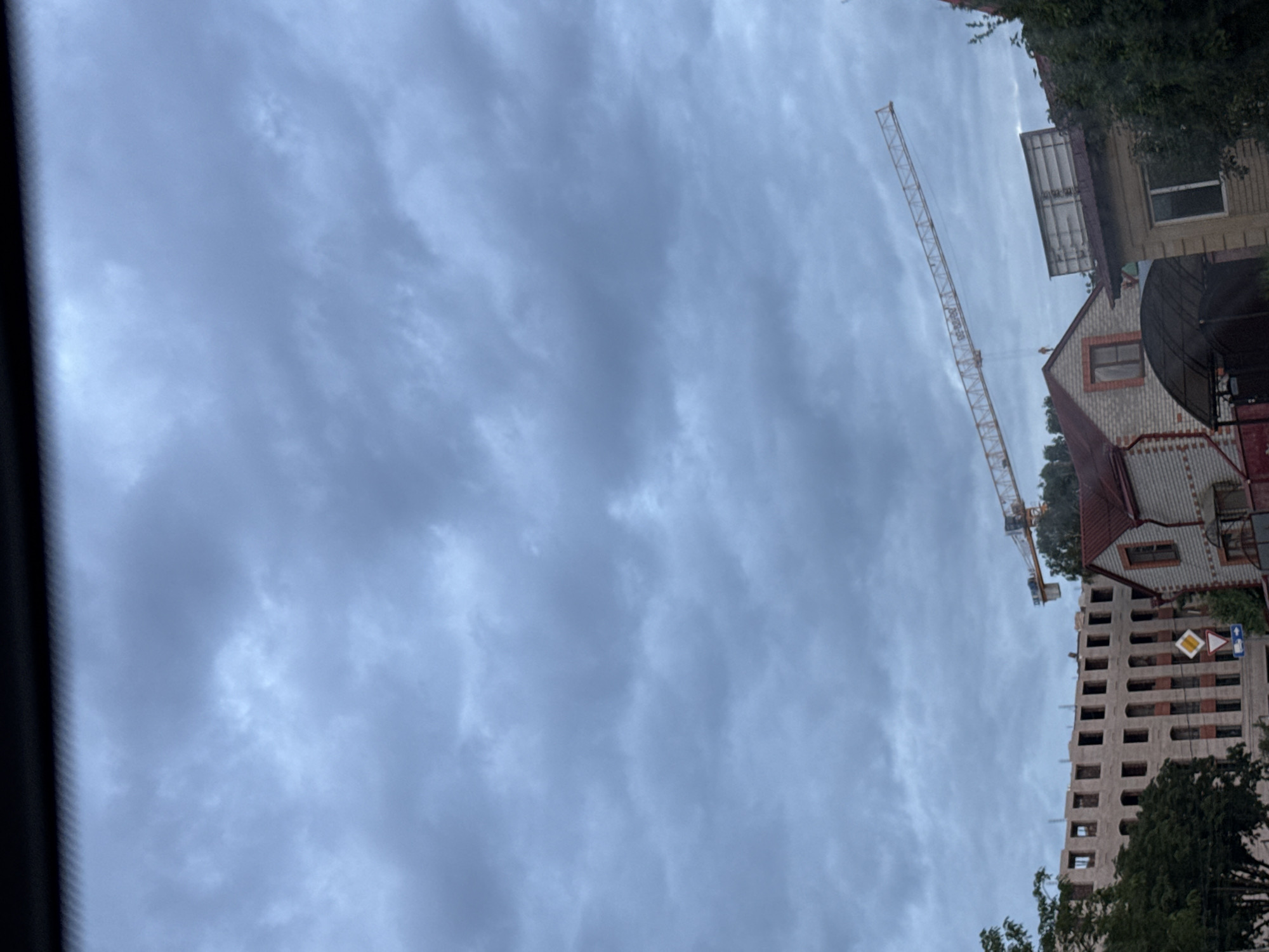 In Mikhaylovsk, Russia, under a cloudy sky, a construction site is visible with a large yellow crane extending diagonally across the frame. To the left, a multi-story building under construction features rows of window openings. In the foreground, two houses with red roofs are present. One house has a gabled roof and visible windows, while the other appears to be a single-story structure. Trees are interspersed throughout the scene, with one prominent tree on the far left.