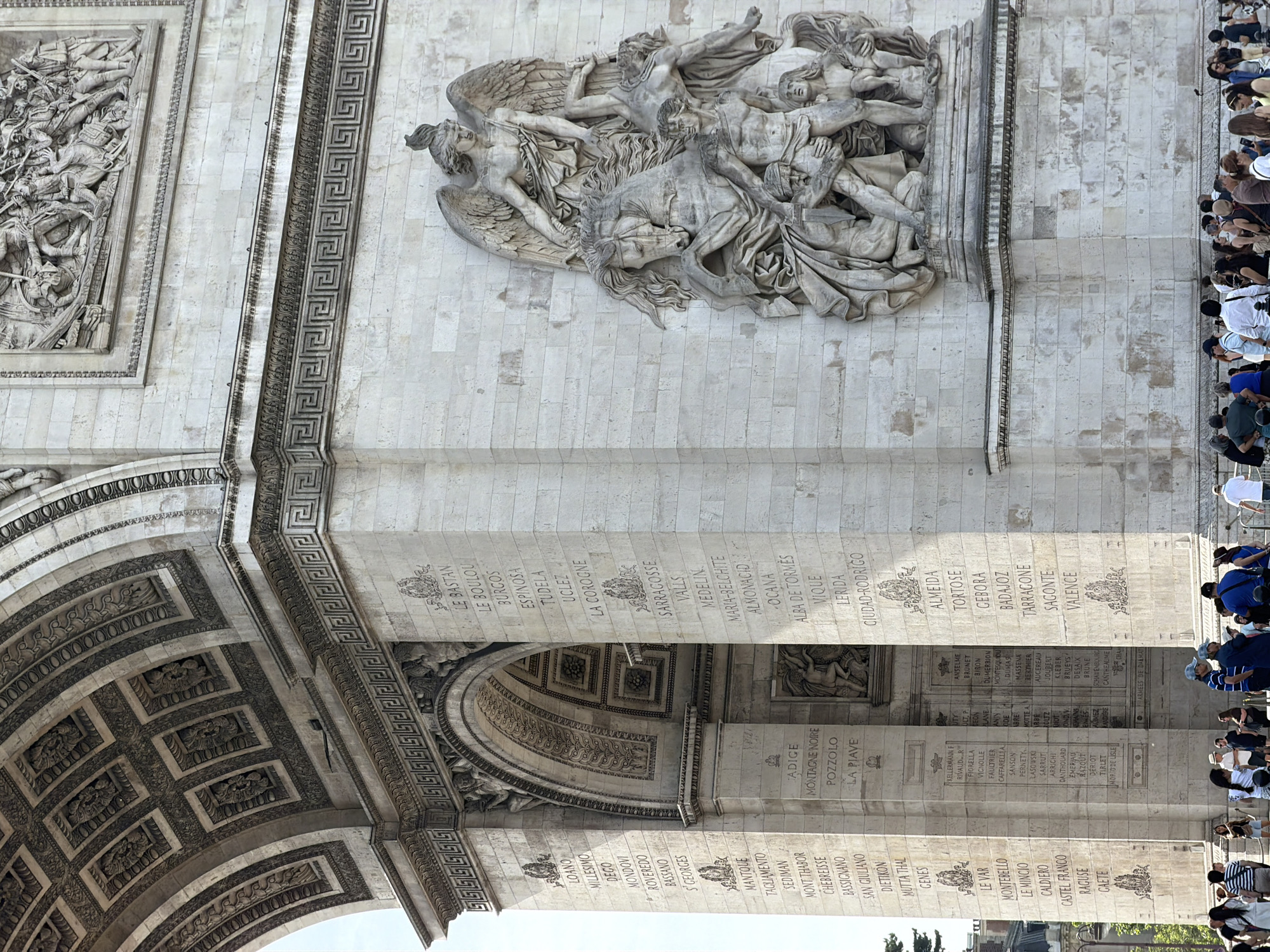 The image shows a portion of the Arc de Triomphe in Paris, France. The structure is made of stone and features architectural details such as carvings, moldings, and inscriptions. Names and places, likely battles or generals, are engraved on the surface. A high-relief sculpture adorns the upper section, depicting multiple figures in a dynamic composition. Along the right edge of the image, a crowd of people is visible, suggesting the structure is a popular tourist destination. The overall lighting suggests a daytime setting.