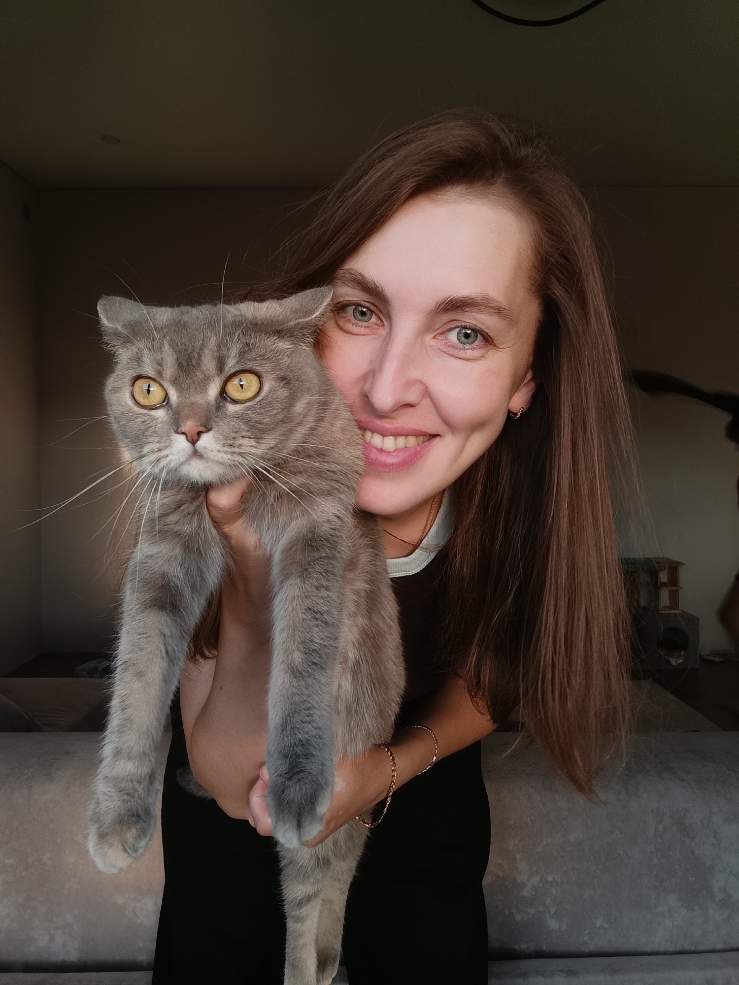 A woman with long brown hair and blue eyes is holding a grey cat. She is smiling at the camera, and the cat is looking at the camera with its mouth open. The woman is wearing a black shirt and a gold bracelet. The cat has yellow eyes and white whiskers. The background is out of focus, but it appears to be an indoor setting with furniture. The lighting suggests it is daytime, possibly late afternoon due to the warm glow. The overall mood is cheerful and affectionate, highlighting the bond between the woman and her pet. No text is visible in the image.