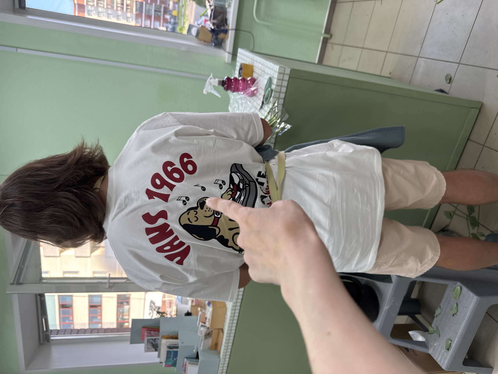 A person wearing a white t-shirt with "VANS 1966" and a cartoon graphic on the back is standing indoors, pointed at by a hand and arm. The person is wearing beige shorts. They are standing near a gray step stool with leaves on it. In the background, there is a green counter with cleaning supplies and a window showing buildings, possibly indicating an urban setting in Izhevsk, Russia. There are also books and bookshelves visible in the lower portion of the window.