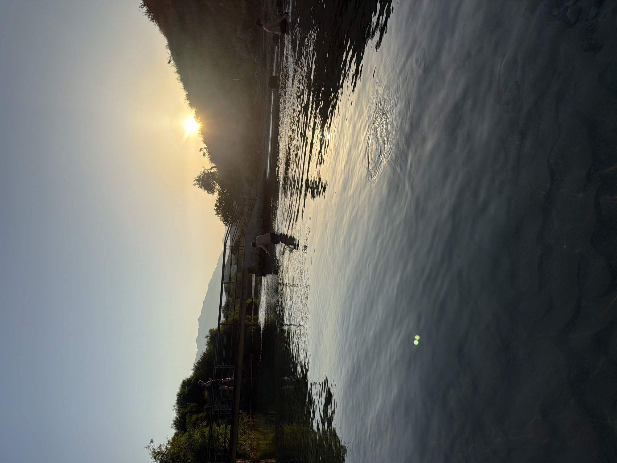 The image depicts a serene natural scene in Georgioupoli, Greece, at sunset. The sun, a bright orb, is setting behind a hill on the right, casting a warm glow across the water. In the foreground, a calm body of water reflects the sky and surrounding landscape, with subtle ripples disturbing its surface. Two bright green circular lights are visible on the water's surface, near the bottom left of the frame.
A pedestrian bridge spans the water in the mid-ground, with two individuals visible on it. One person, appearing to be walking, is on the left side of the bridge. Another person is standing in the water, wading near the bridge's support structure. Further to the right, two more individuals are present in the shallow water. One is partially submerged and appears to be sitting or kneeling, while another person is visible further back in the water.
The background features lush green vegetation and the silhouette of mountains against the clear, pale blue sky. The overall atmosphere is tranquil, capturing the peaceful ambiance of a sunset by the water.