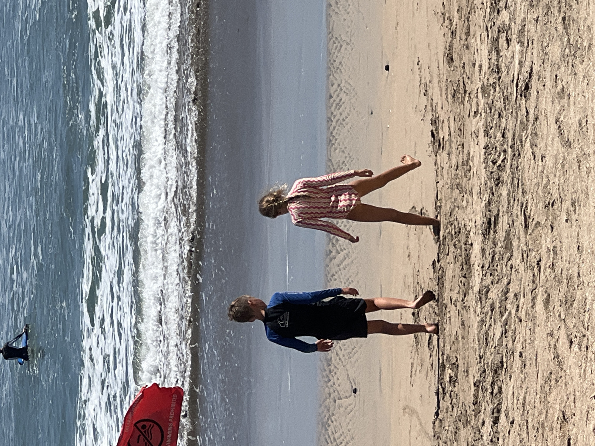 The image depicts a beach scene in Kuta, Indonesia. In the foreground, two children, a boy and a girl, are standing on the wet sand facing the ocean. The boy, wearing a blue and black long-sleeved rash guard and black shorts, is on the left. The girl, on the right, is wearing a pink and white striped one-piece swimsuit. Her hair is in a braid.
In the middle ground, the ocean waves are breaking on the shore. A red flag with a white symbol is planted in the sand near the water's edge, with the text "SWIMMING PROHIBITED" visible. In the background, further out in the ocean, a person is sitting on a surfboard. The water is a deep blue with white foam from the waves. The sand is a light brown color, showing evidence of being recently wet. The overall lighting suggests a sunny day.