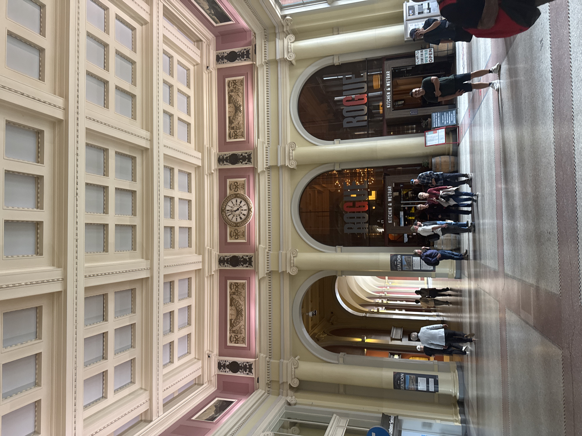 The image shows the interior of a train station in Vancouver, Canada. The ceiling is highly ornate with a coffered design and decorative molding. The walls are painted a pale yellow with a pink band near the ceiling, which features decorative panels and a large clock. Several large arched openings lead to other parts of the station, with one on the right displaying the signage "ROGUE KITCHEN & WETBAR."
In the foreground, a man in a dark green t-shirt and black shorts stands looking towards the camera. To his left, a group of people are walking through the station, including a man in a plaid shirt and jeans, and a woman in a red top. To the far right, a person wearing a black t-shirt and cap is seen with their hand to their face. Further back, several other individuals are walking in various directions. The floor is tiled in a geometric pattern. Signs for "THE STATION" are visible on pillars. The overall scene depicts a busy transit hub with architectural grandeur.