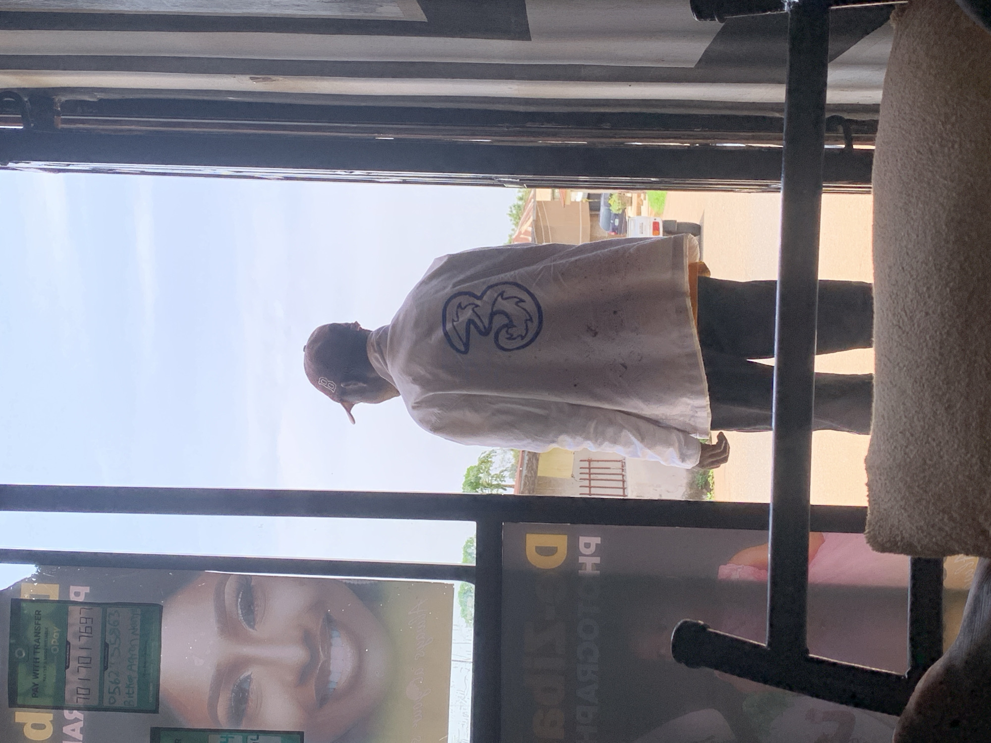 The image captures a candid daytime scene, viewed from inside a building looking out through a window or doorway. The primary subject is a person, seen from behind, standing and looking towards the outside. They are wearing a light-colored, possibly off-white, long-sleeved shirt or jacket, prominently featuring a dark blue "3" logo on the back. A dark baseball cap is also visible on their head, and dark trousers complete their attire.
In the immediate foreground, to the left, there's a large poster or screen displaying a smiling woman's face. Below this, payment information is visible, including "PAY WITH TRANSFER" and the platform "OPay." Several numerical sequences, likely phone numbers or account details, are present: "054210101697" and "0562755863." There are also fragmented words such as "KAN TER A MORORITJABLK" and "Bitka Agron Narys," which appear to be instructions or details related to the payment. To the right, a textured, light-colored object, possibly a piece of furniture like a fabric-covered armrest or cushion, is partially visible, along with a dark metal frame structure that divides the view.
Beyond the person and the interior elements, the background reveals an outdoor setting under a bright, somewhat hazy sky, indicating daytime. Buildings, possibly residential structures, with walls and fencing, are visible. Trees add greenery to the distant scene. A white vehicle, possibly a van or truck, and another darker vehicle can be seen parked or driving on what appears to be a street or open area. The architecture and the payment details including OPay and local phone prefixes are consistent with the provided context of Jos, Nigeria.
