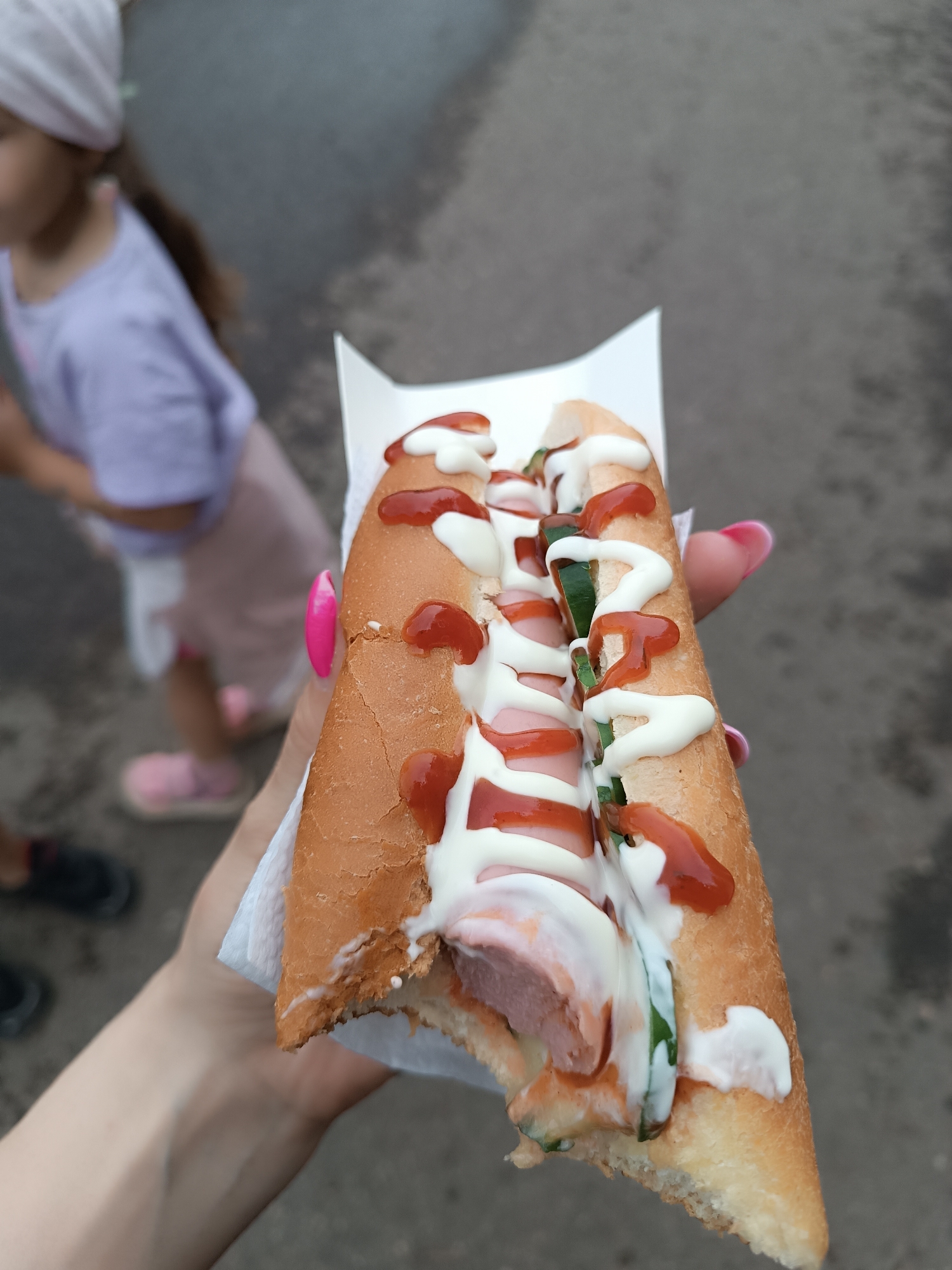 A person is holding a partially eaten hot dog in a white cardboard container. The hot dog has ketchup, mayonnaise, and green strips, possibly cucumber, on top. The person holding the hot dog has pink nail polish. In the background, a child in a light purple shirt and pink shorts and shoes is partially visible, along with a glimpse of shoes belonging to another individual. The ground appears to be a paved surface. There is no information about the location of the image.