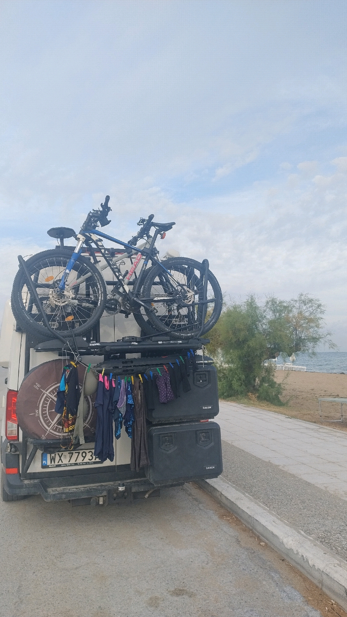 The image shows the rear of a white van parked on a paved surface, possibly a road or parking area. The van is equipped with a roof rack carrying two bicycles. Below the bicycles, a rack holds a collection of drying laundry, secured with clothespins. The laundry includes various items of clothing in different colors and patterns. Two black storage boxes, labeled "DAKEN," are attached to the rear of the van. A spare tire cover with a compass design is mounted on the left rear of the van. The license plate reads "WX 7793".
The background reveals a beach scene with a body of water, likely the sea, and a sandy shore. There are trees and some white structures, possibly beach chairs or umbrellas, visible in the distance. The sky is overcast with clouds. The overall setting suggests a coastal location, potentially a campsite or a parking area near a beach.
The location of Neoi Epivates, Greece, is identified by the license plate, which includes "PL" indicating Poland, but the context provided by the user links it to this specific Greek location. This suggests the vehicle might be a tourist's vehicle in Greece.
There are no people visible in the image. The van appears to be stationary. The environment is a mix of urban infrastructure (paved surface, van) and natural elements (sea, beach, trees). The lighting suggests daylight, possibly morning or late afternoon, given the diffused light and the overcast sky.