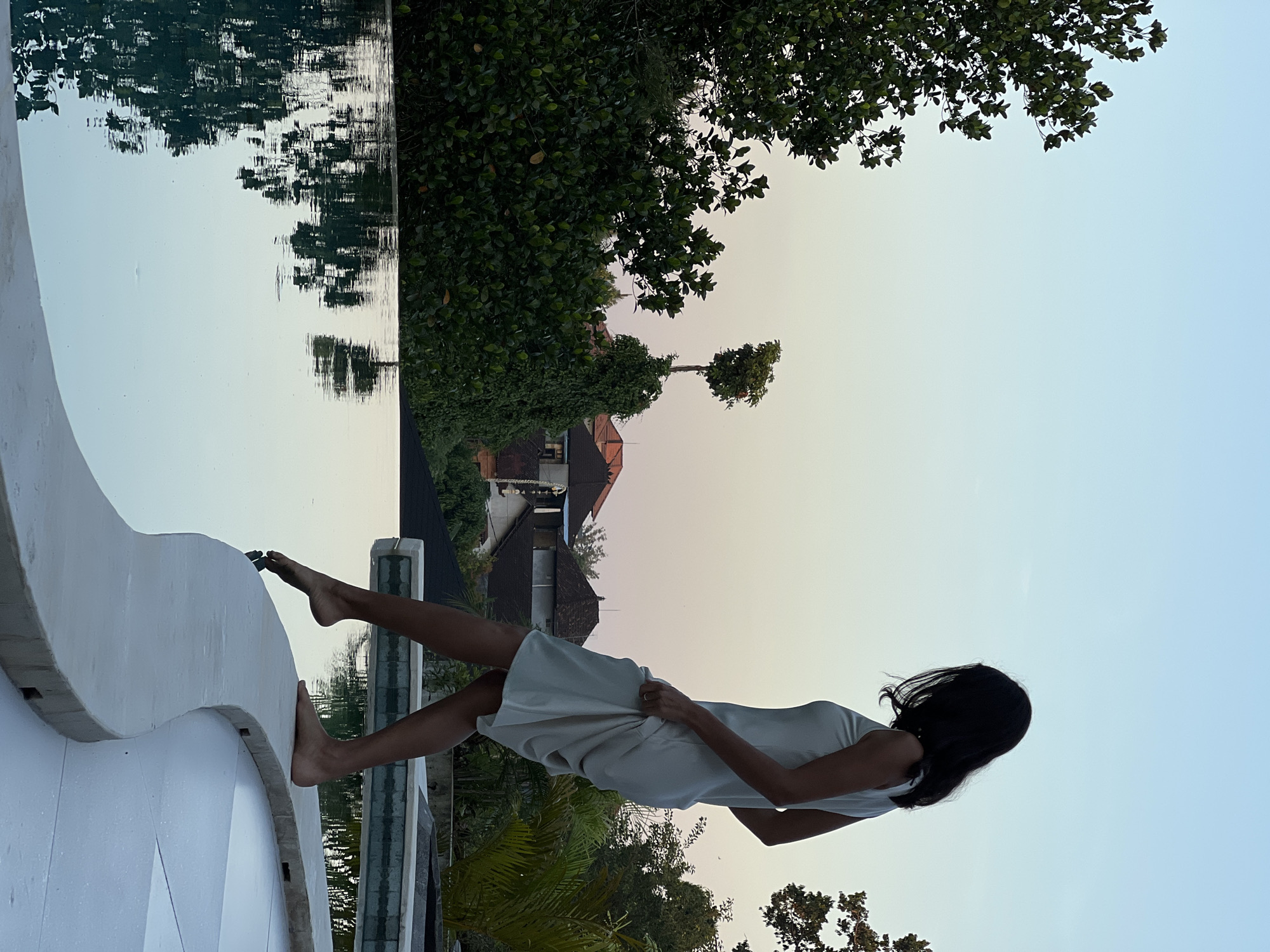 A woman stands on the edge of an infinity pool in what appears to be a resort or private residence in Ubud District, Indonesia. She is wearing a light-colored, sleeveless dress and is silhouetted against a pale sky. Her right foot is extended towards the water, with her toes just touching the surface, creating ripples. The pool reflects the surrounding lush green foliage and the soft hues of the twilight sky. In the background, traditional Indonesian architecture with tiled roofs is visible amidst the dense vegetation. The scene conveys a sense of tranquility and evening relaxation.