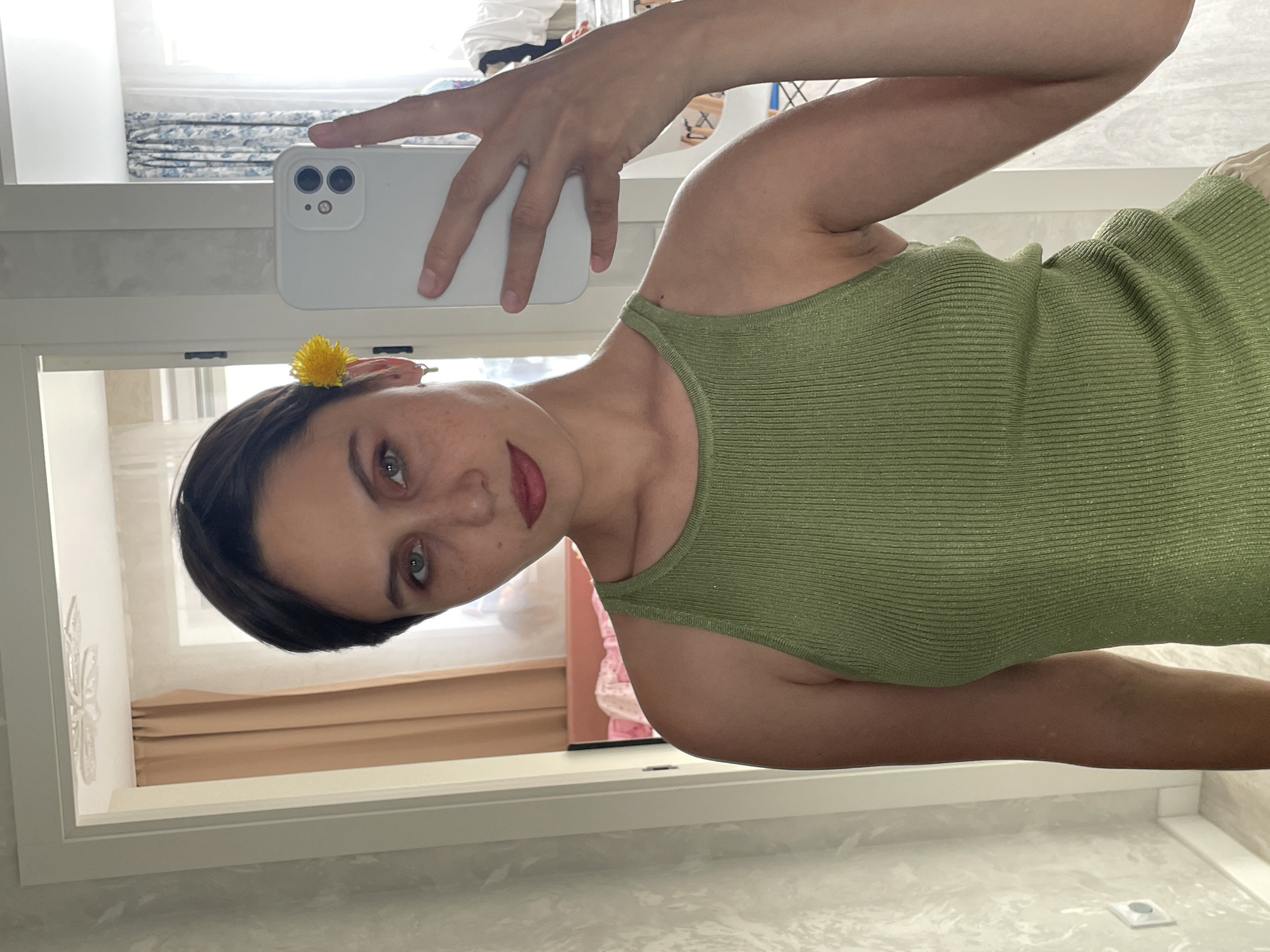 A woman is taking a selfie in what appears to be a bathroom or bedroom. She's holding a white smartphone in her right hand. She's wearing a green tank top and has a small yellow flower tucked behind her ear. The background shows a window with curtains, and a partially visible mirror reflects another room. The location cannot be determined as Izhevsk, Russia based on the available image information.