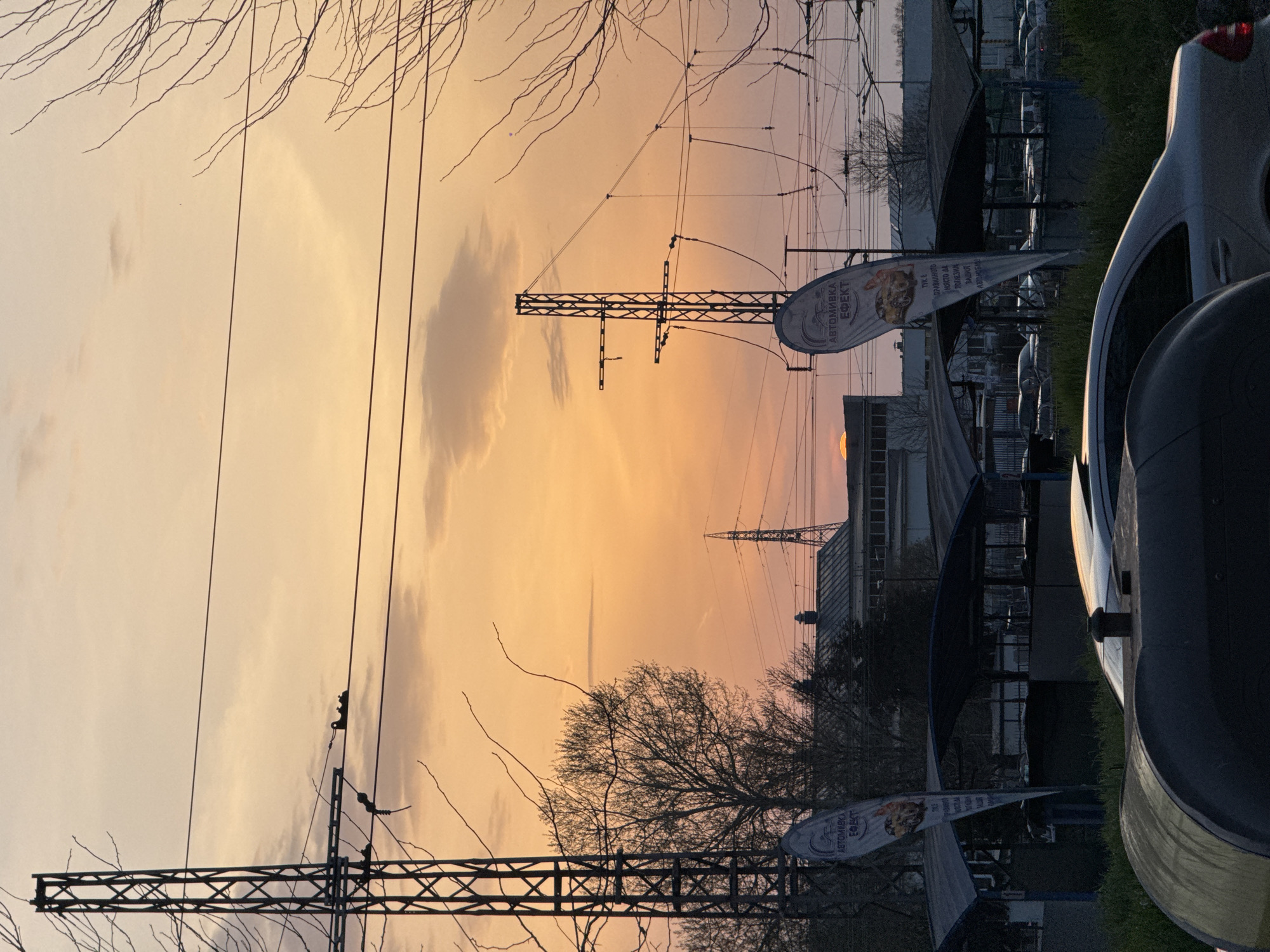 The image shows a sunset in what appears to be an urban or suburban area of Burgas, Bulgaria, based on the Cyrillic text on a nearby flag or banner. The sky is predominantly orange and yellow, suggesting either sunrise or sunset. Power lines and utility poles are visible against the sky. Bare tree branches are silhouetted in the foreground. A building is visible in the midground. In the extreme foreground, a parked car and what appears to be a trash can are seen.